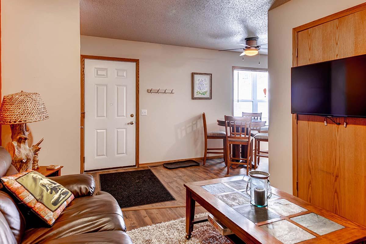 A cozy living room with a brown leather sofa, a coffee table with a decorative centerpiece, and a flat-screen TV mounted on the wall. A door leads to the outside, and in the background, a dining area with a wooden table and chairs is visible. The space is well-lit and inviting.
