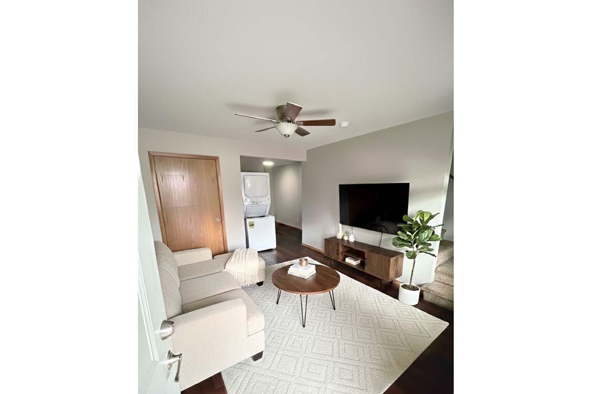 A cozy living area featuring a light-colored sofa set, a round wooden coffee table, and a potted plant. The room includes a modern flat-screen TV mounted on the wall and a doorway leading to a laundry area. Natural light brightens the space, complemented by neutral wall colors and a textured area rug.