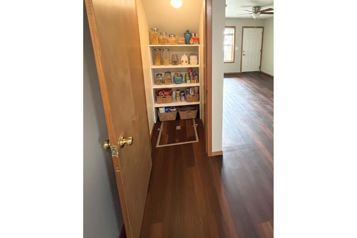 A view of a pantry entrance with a wooden door partially open, revealing organized shelves filled with various jars and containers. The floor is dark brown, and in the background, there's a spacious room with a ceiling fan and doors leading outside. Natural light fills the space, creating a warm ambiance.