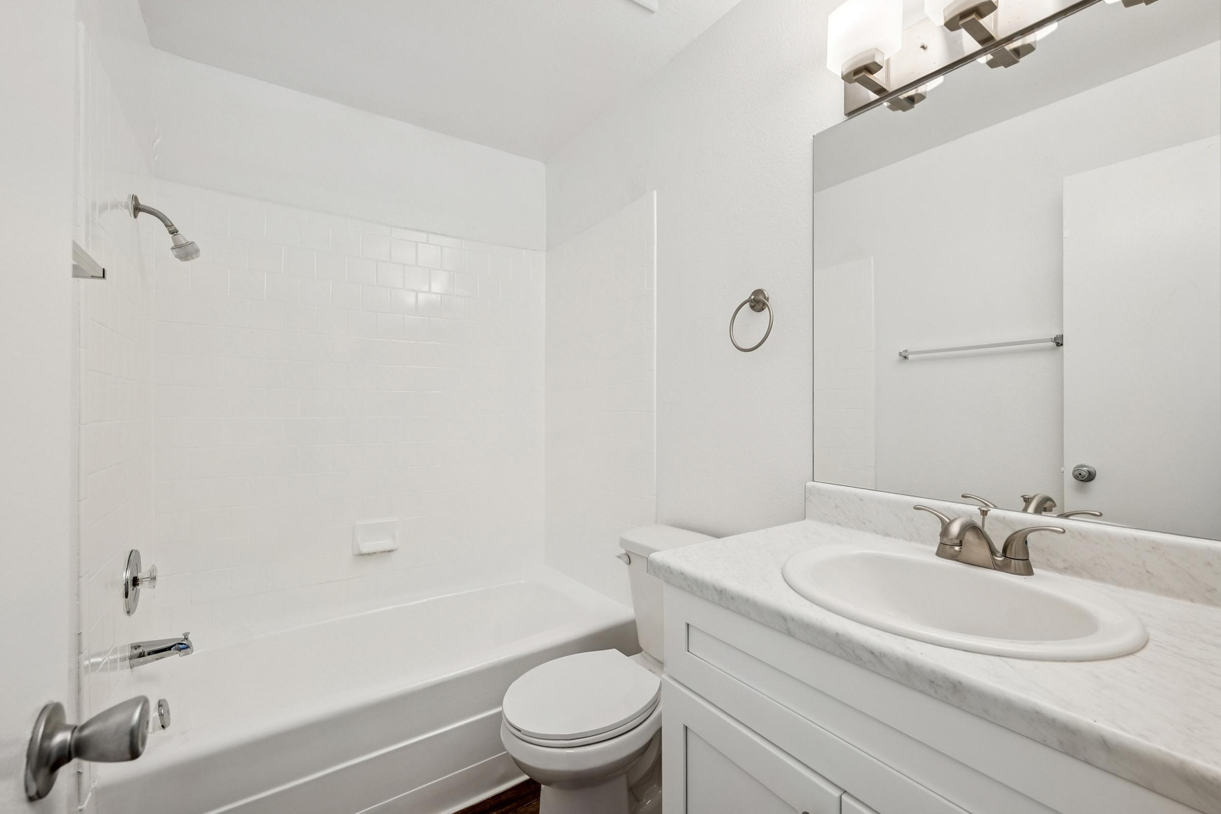 A clean, modern bathroom featuring a bathtub with a showerhead, a white toilet, and a vanity with a marble countertop and sink. The walls and shower area are tiled in white, and there are two light fixtures above a large mirror. The floor has dark wood accents.