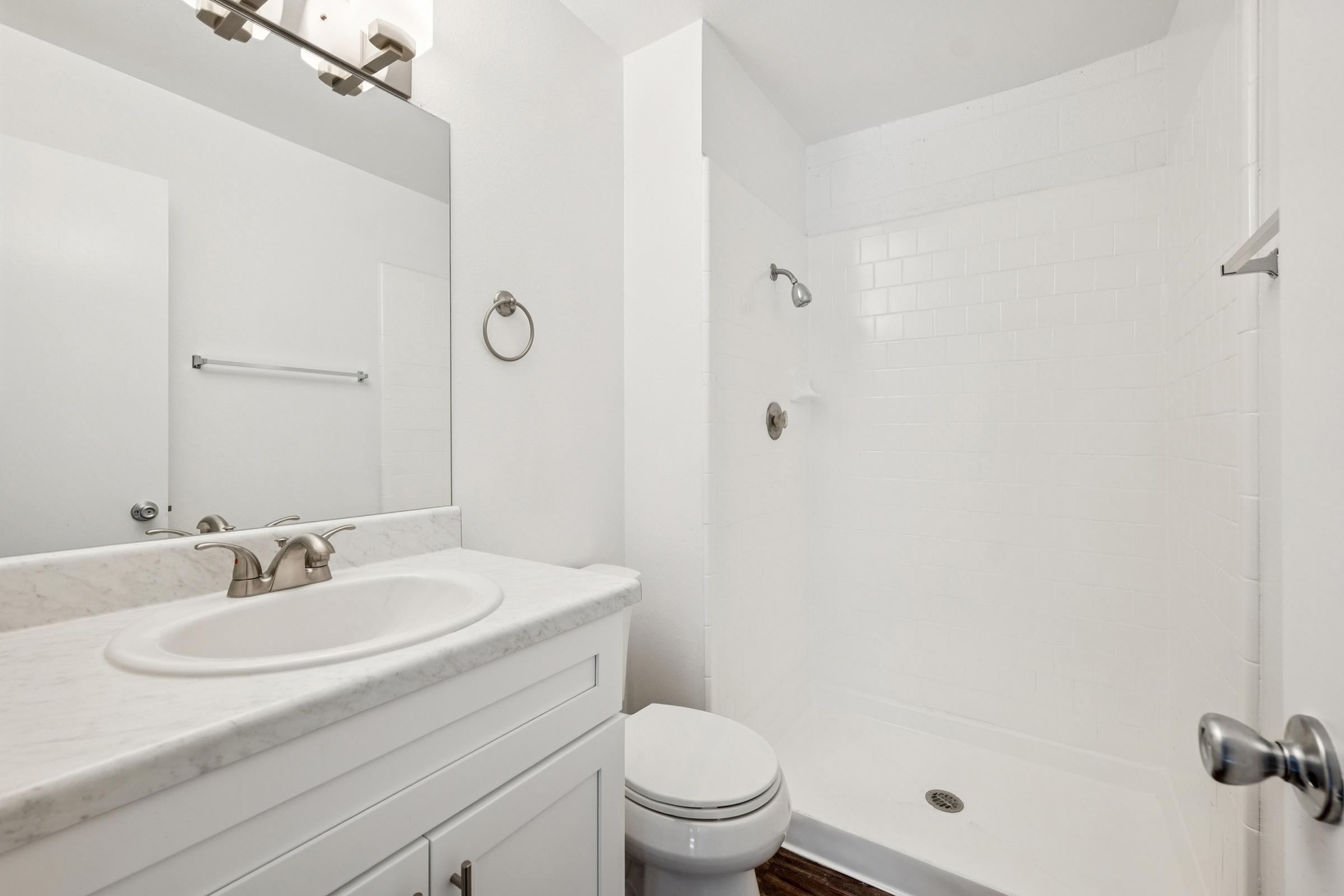 A clean, modern bathroom featuring a white tiled shower, a white vanity with a marble countertop, a round mirror above the sink, a toilet, and a chrome towel ring. The walls are painted white, creating a bright and spacious appearance.
