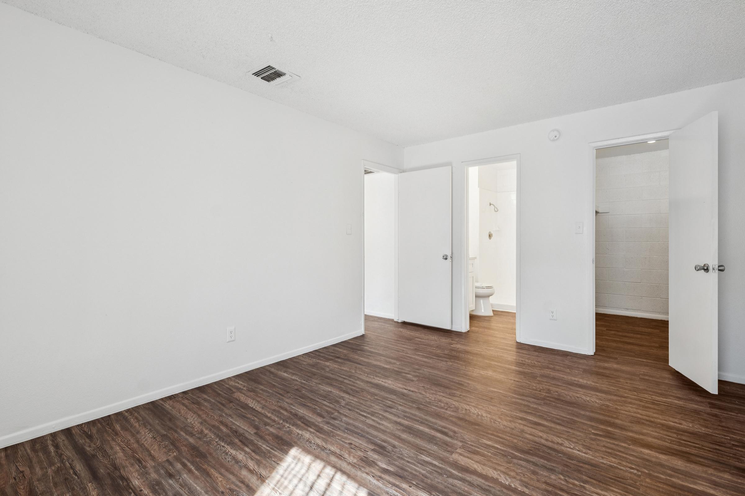 Interior view of a vacant room with light-colored walls and a wood-like floor. Two doors are visible, one leading to a bathroom and the other to another room. The space is well-lit, with natural light coming from a window, creating a clean and open atmosphere.