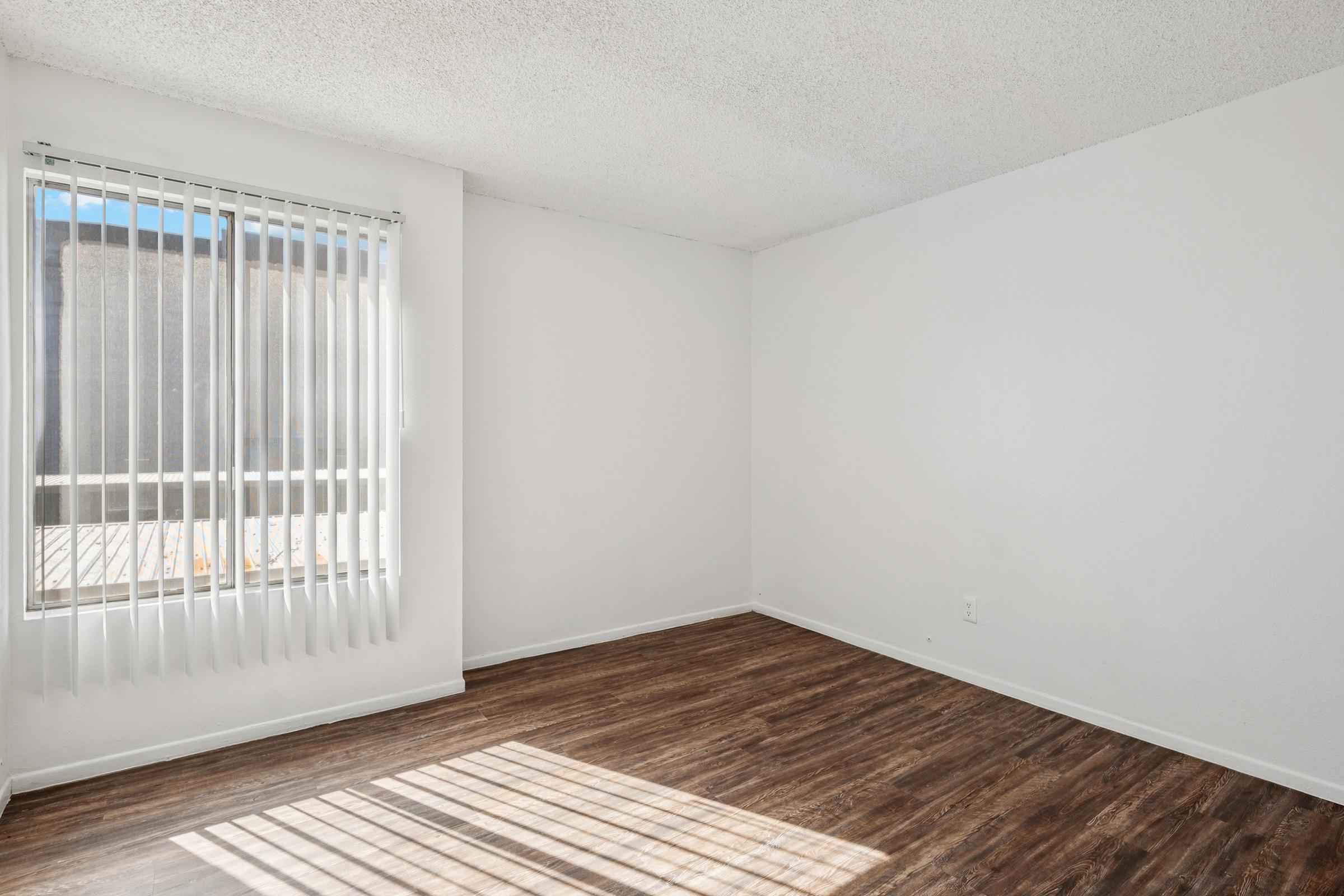 A blank room with light-colored walls and a wooden floor. There are vertical blinds on a window allowing natural light to enter. The space is empty, creating a minimalistic and clean appearance, with no furniture or decorations.