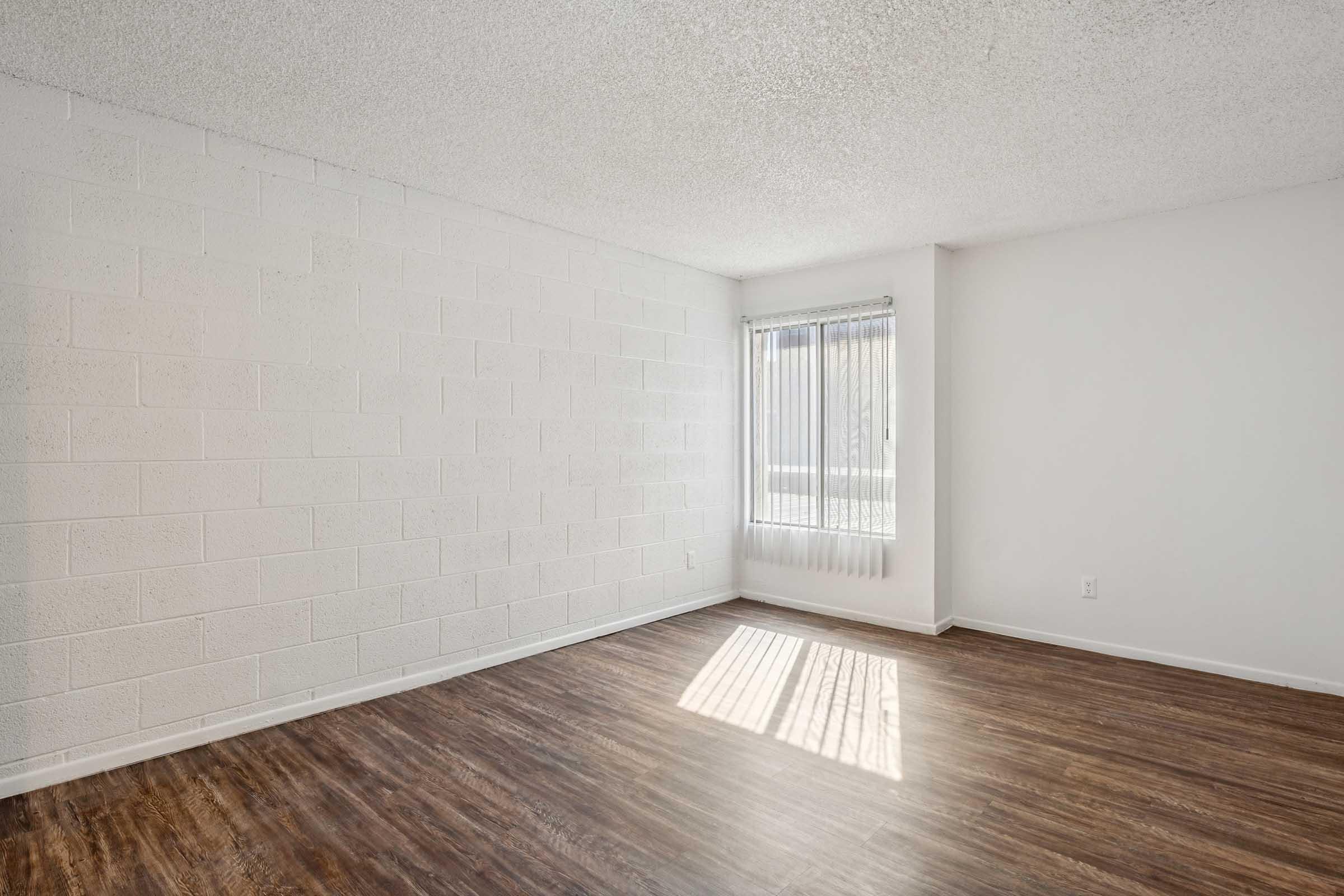 A spacious, empty room featuring a textured white wall, a large window with vertical blinds allowing natural light, and dark wood-like flooring. The room appears bright and minimalistic, providing a blank canvas for decoration.