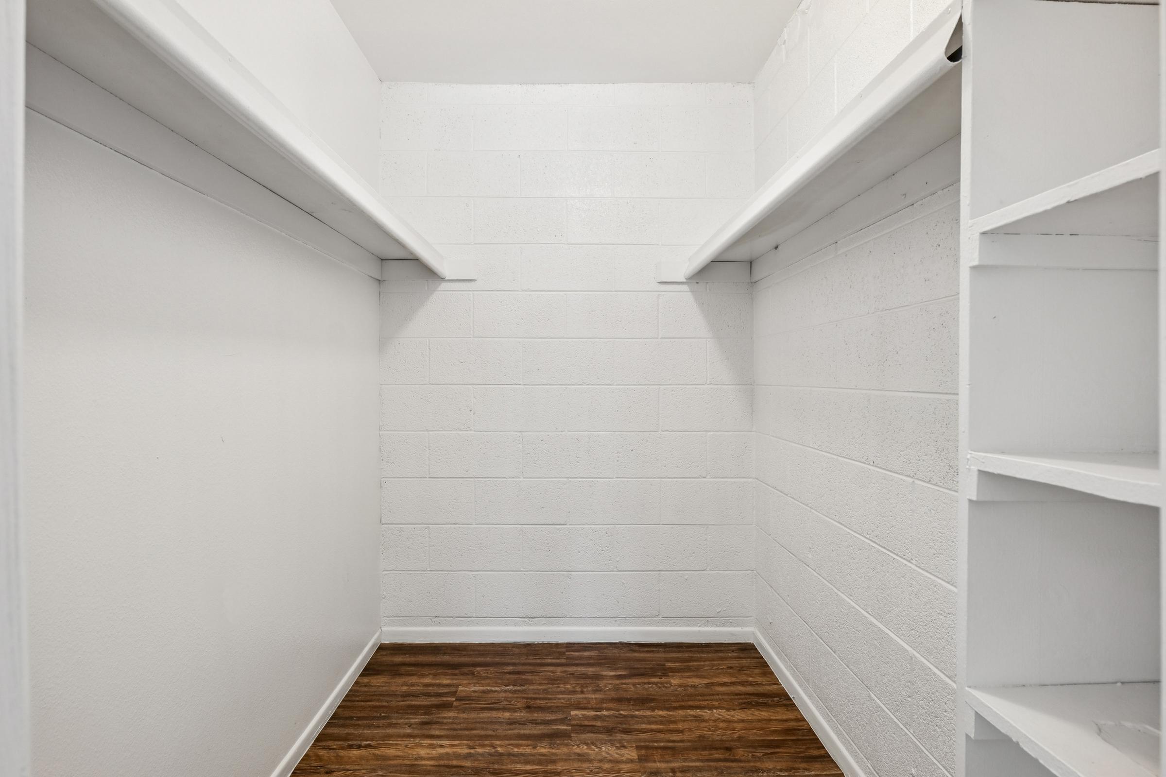 Empty closet with white cinder block walls and two shelves on either side. The floor features a wooden finish, creating a spacious and minimalistic storage area.