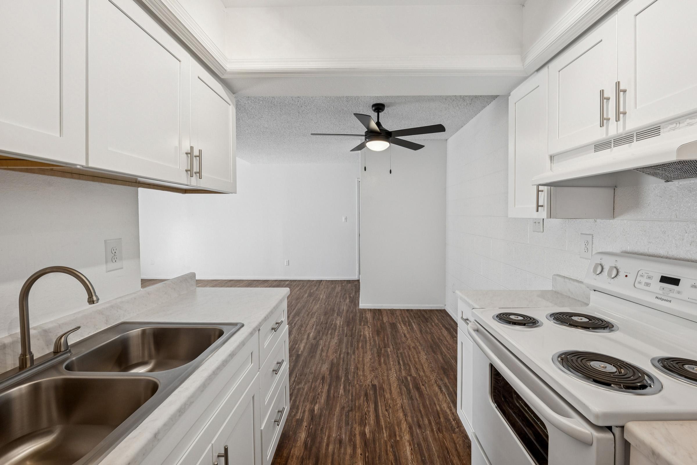 Modern kitchen featuring a double sink with a brushed nickel faucet, white cabinetry, granite countertops, and a black ceiling fan. An electric stove and oven are visible, along with a microwave above. The open layout leads to a spacious living area with wooden flooring and light-colored walls.