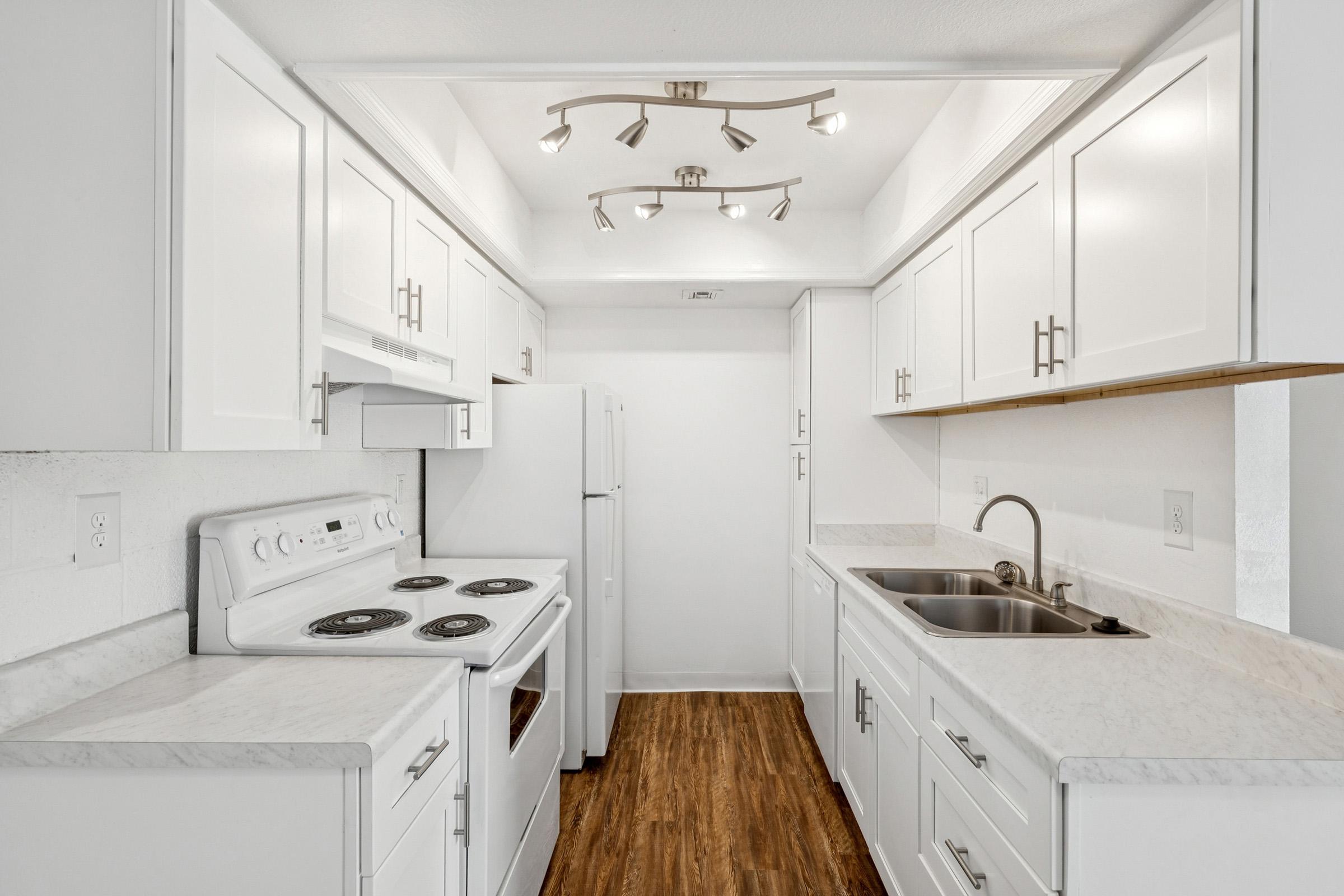 A modern kitchen with white cabinets, a white stove, and a double sink. The countertops are light-colored with a marble pattern, and there's a refrigerator visible. The ceiling features track lighting, and the flooring is a warm wood finish, creating a bright and inviting atmosphere.