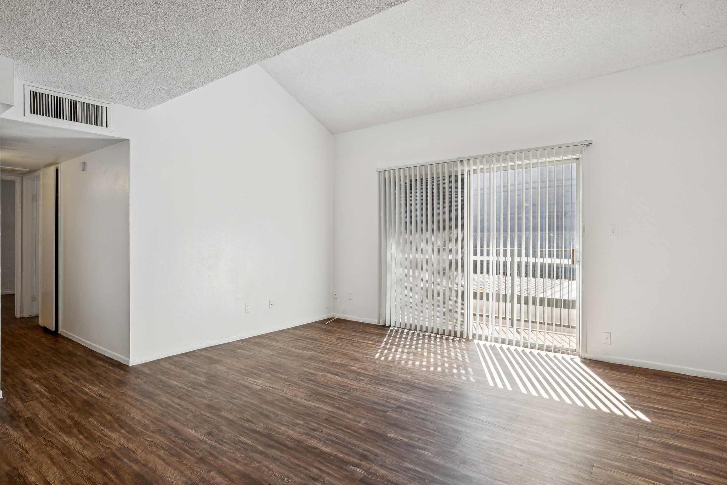 A spacious, well-lit living room with a high ceiling, featuring light-colored walls and dark wood-style flooring. There is a sliding glass door with vertical blinds leading to a balcony, allowing natural light to enter the room. The area appears clean and ready for furnishing.