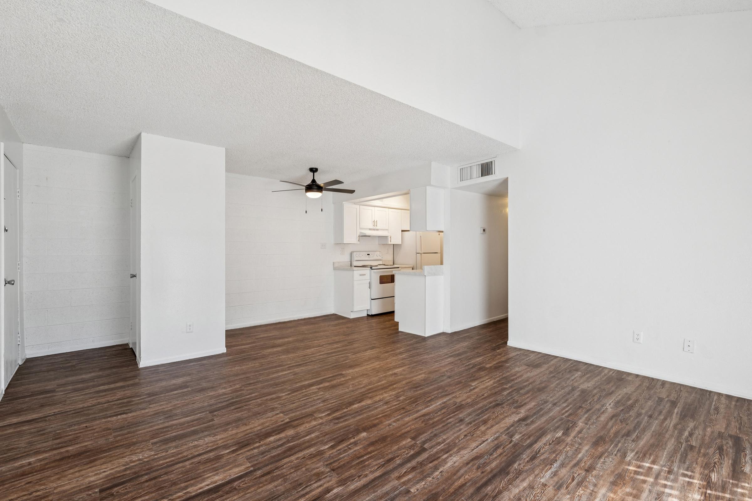 Modern, open-concept living space featuring light wood-like flooring, white walls, and a ceiling fan. To the left, there is a doorway leading to another room, and the kitchen area is partially visible in the back, with white cabinets and appliances. Natural light fills the area, creating a bright atmosphere.
