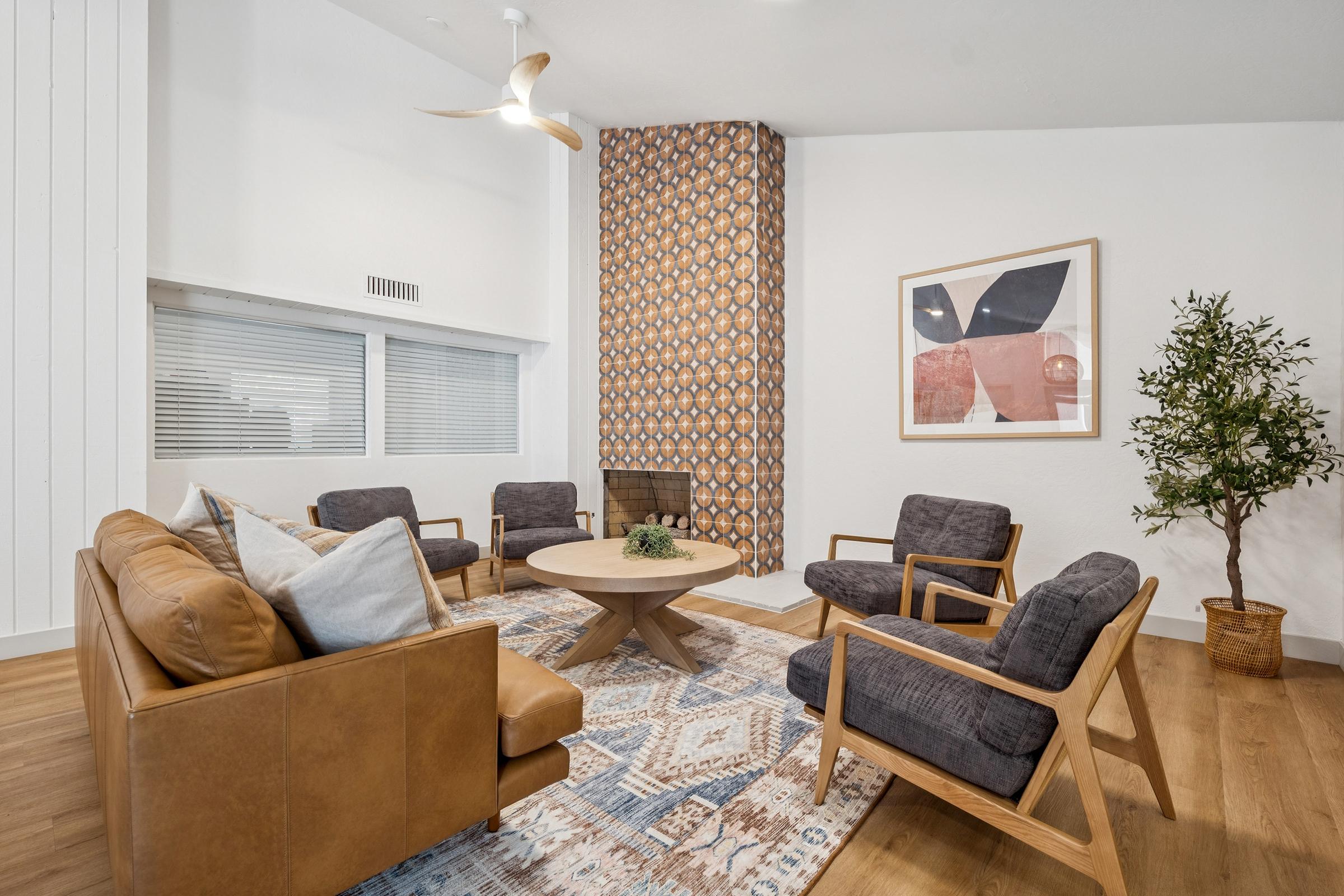 A cozy living room featuring a modern design with a brown leather sofa, four wooden chairs with dark cushions, and a round coffee table. The space includes a patterned accent wall, a fireplace, and a decorative plant. Natural light fills the room through large windows, creating an inviting atmosphere.