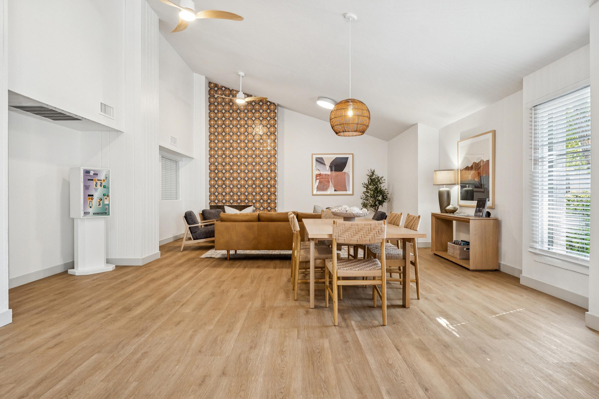 A modern, open living area featuring a dining table with wooden chairs, a couch set against a textured accent wall, a television unit, and decorative elements. Large windows provide natural light, and a ceiling fan hangs above. The flooring is light wood, creating a warm and inviting atmosphere.