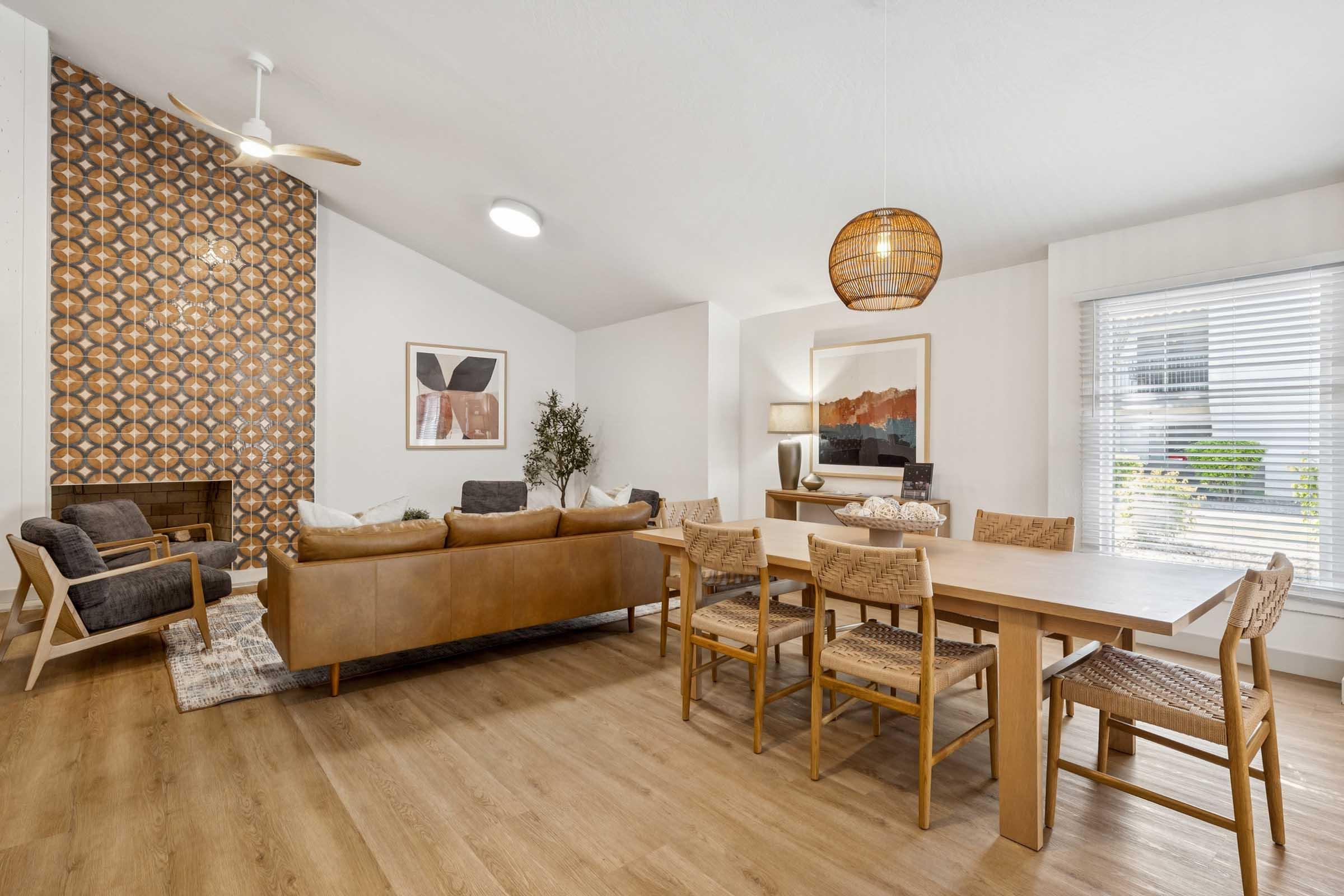 A modern living and dining area featuring a brown leather sofa, wooden dining table with woven chairs, and stylish decor. The space includes a textured accent wall, light wood flooring, a ceiling fan, and large windows allowing natural light in, creating an inviting atmosphere.