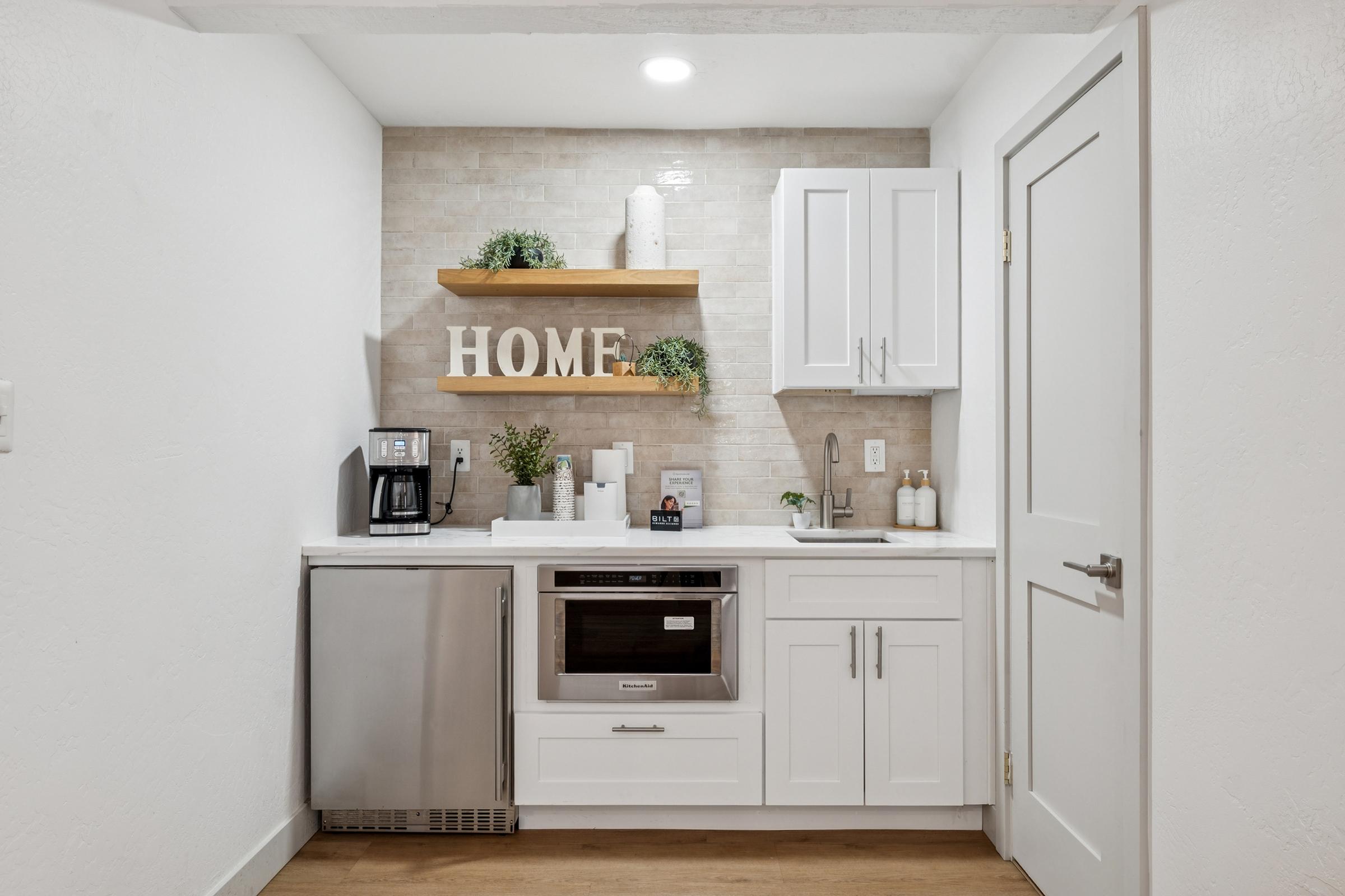 A modern kitchen corner featuring white cabinetry, a stainless steel refrigerator, and a built-in microwave. Decor includes a wooden shelf with small plants and a decoration spelling "HOME." The countertop is clean and organized, with a coffee maker and various containers arranged neatly.