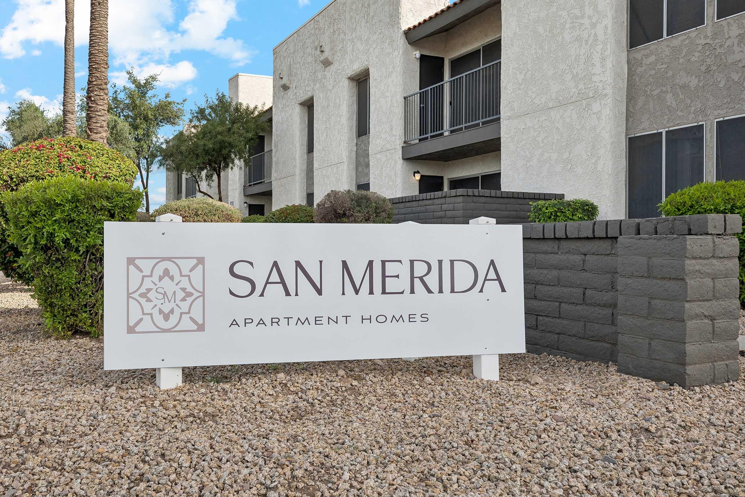 Sign for San Merida Apartment Homes, featuring the name prominently displayed with a decorative logo. The sign is situated in front of a landscaped area with shrubs and decorative rocks, alongside multi-story apartment buildings in the background. Blue sky and palm trees are visible.
