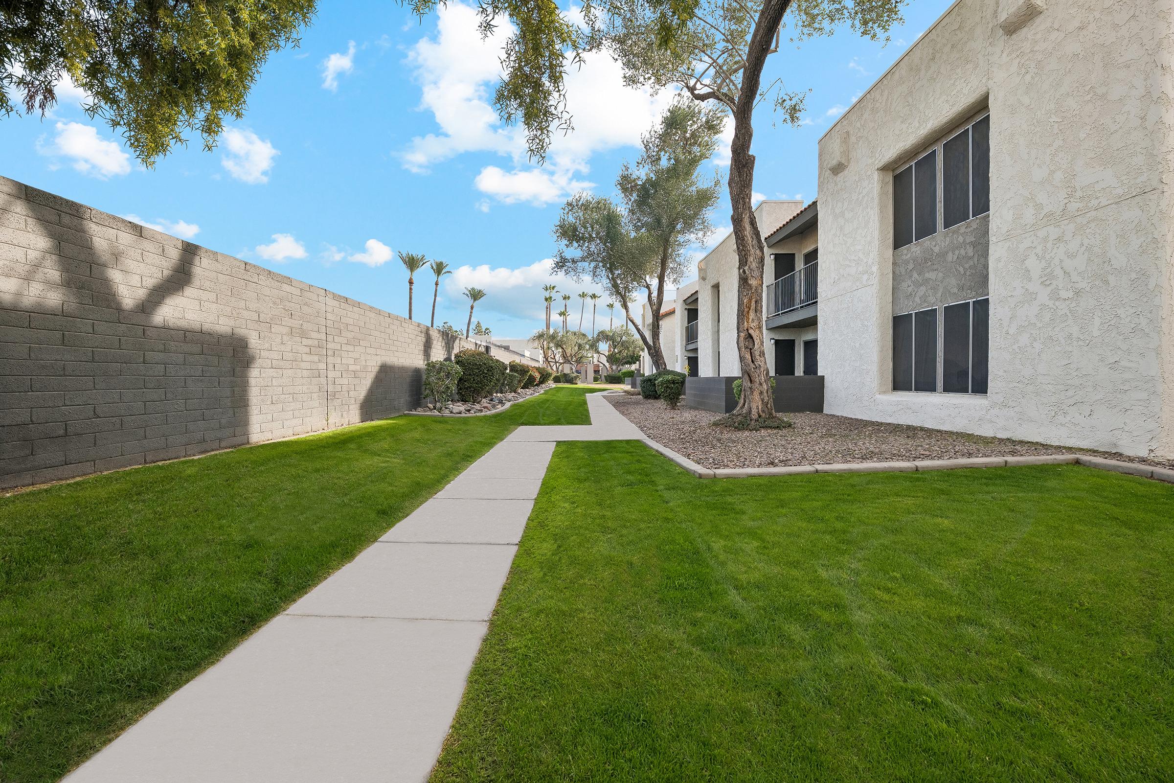 A well-maintained pathway lined with grass runs between two buildings, leading towards a landscape featuring palm trees. The sky is bright with scattered clouds, and a gray wall borders the left side of the scene, enhancing the serene outdoor environment.