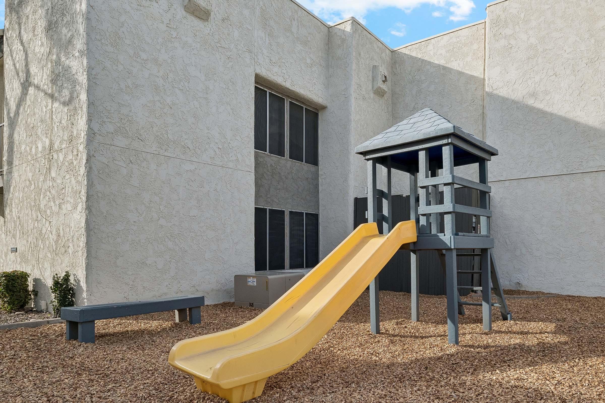 Playground area featuring a wooden play structure with a yellow slide, surrounded by a gravel surface. In the background, there are large windows of a building, and a small bench nearby. The setting is bright and open, ideal for children to play.