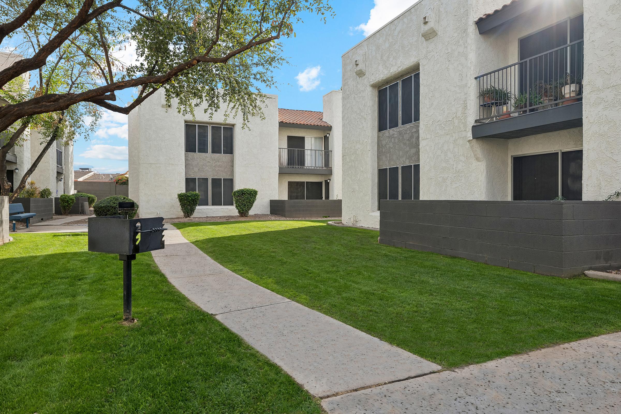 A view of a well-maintained residential apartment complex with two white buildings featuring dark gray accents. There is a manicured lawn with a pathway leading through the greenery, bordered by low hedges and potted plants on balconies. A mailbox stands near the walkway, adding to the serene atmosphere.