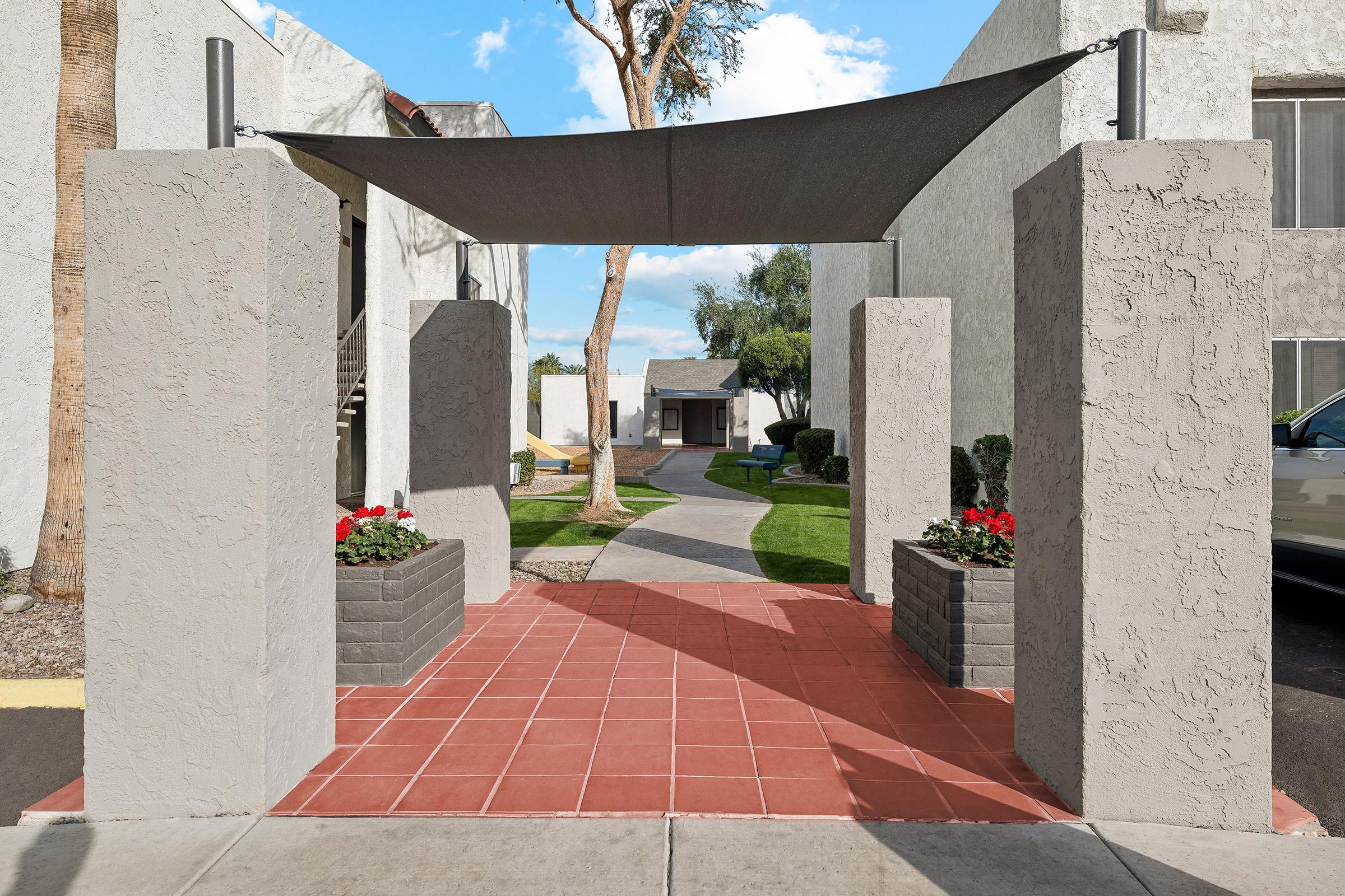 A walkway leading through a shaded entrance with grey pillars and planters containing red flowers. The path is surrounded by green grass and trees, with buildings in the background under a blue sky with clouds.