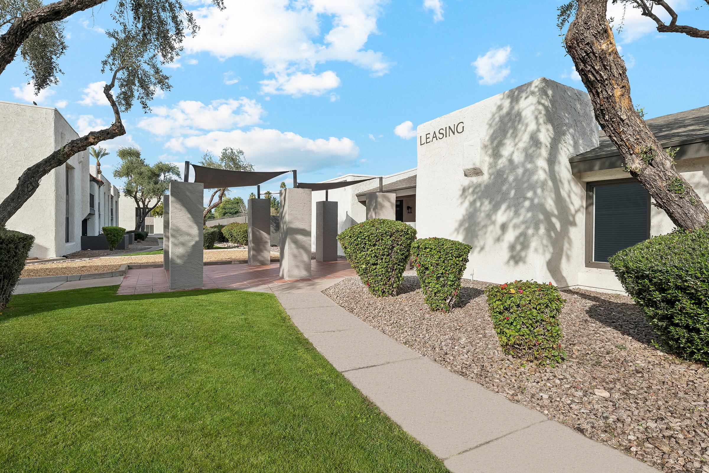 A view of a leasing office area featuring modern architecture. The path leads to a building labeled "LEASING," surrounded by neatly trimmed bushes and trees. Lush green grass and a sunny blue sky enhance the inviting atmosphere of the property.