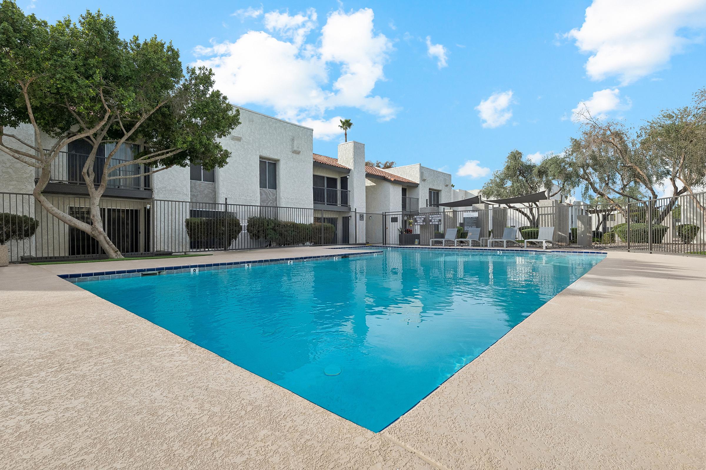 A clear swimming pool with a blue surface, surrounded by a concrete deck. In the background, there are several two-story apartment buildings, along with well-maintained trees and shrubs. The sky is bright and partly cloudy, creating a serene outdoor atmosphere.
