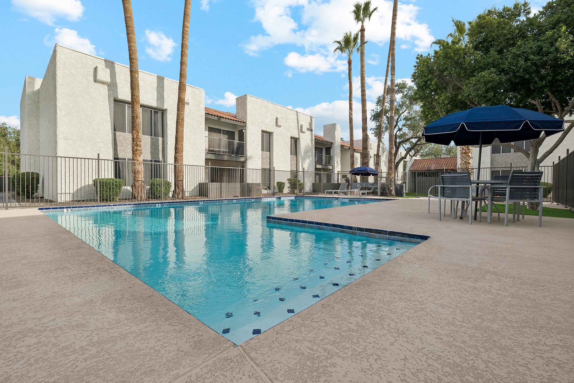A sunny community pool area featuring clear blue water, surrounded by palm trees and lounge chairs. In the background, there are apartment buildings with balconies. A large blue umbrella provides shade near a seating area with tables and chairs beside the pool. The sky is partly cloudy.