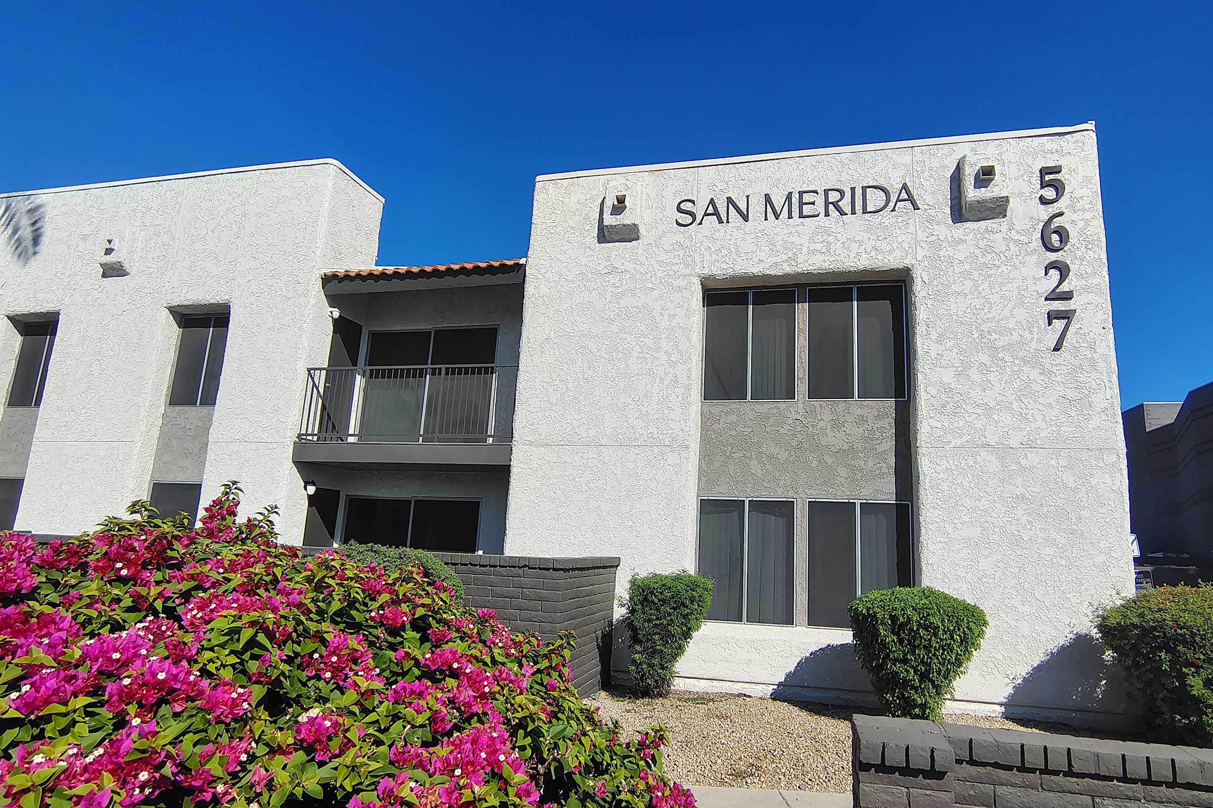 A two-story residential building with a textured white exterior. The words "SAN MERIDA" are prominently displayed on the front, above the entrance. The building features multiple windows and a balcony. Colorful bougainvillea plants are in the foreground, adding vibrancy to the scene against a clear blue sky.