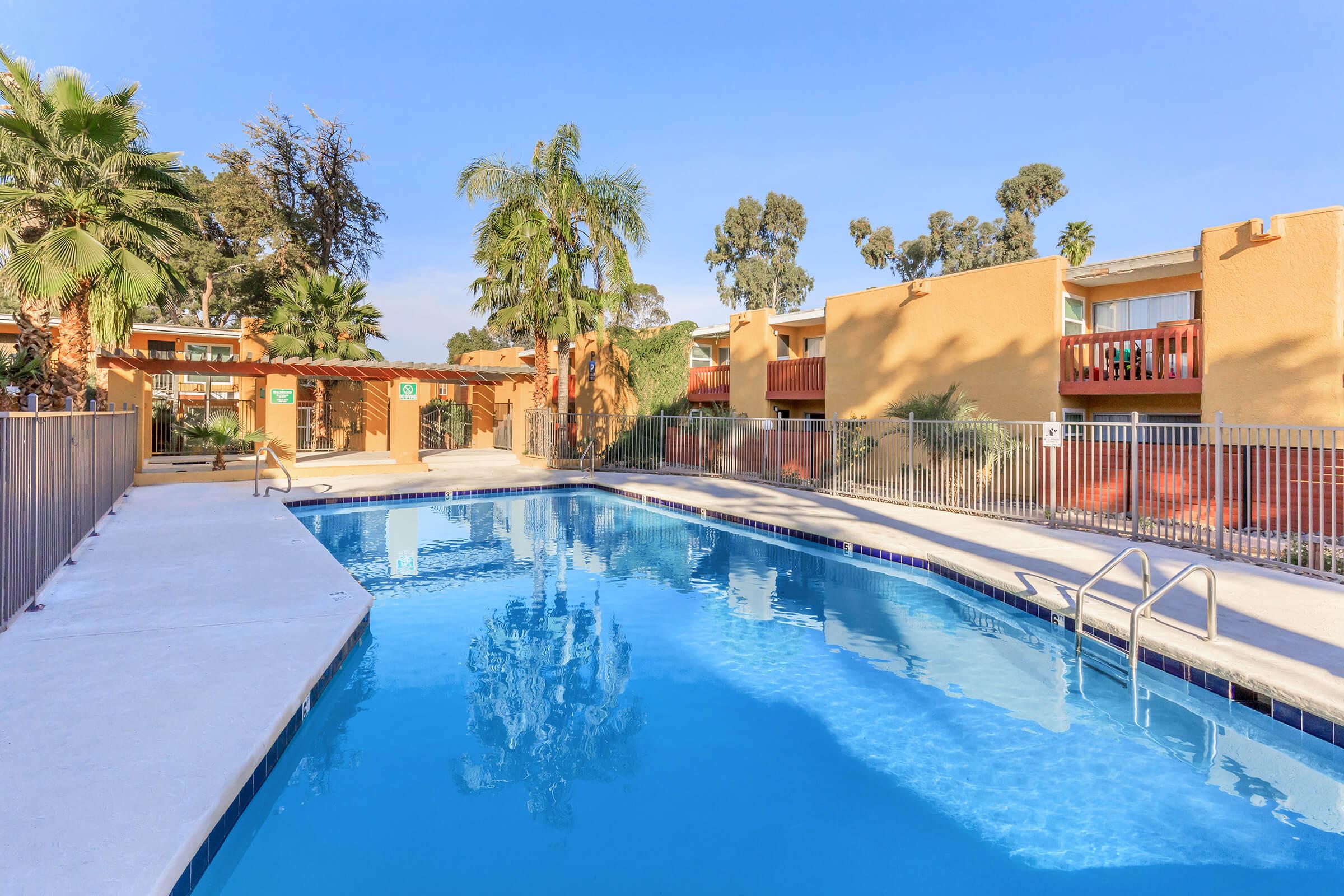 A clear blue swimming pool surrounded by palm trees and greenery, with lounge chairs nearby. In the background, orange-colored apartment buildings with balconies are partially visible. A fence encloses the pool area, and the sky is bright and sunny.