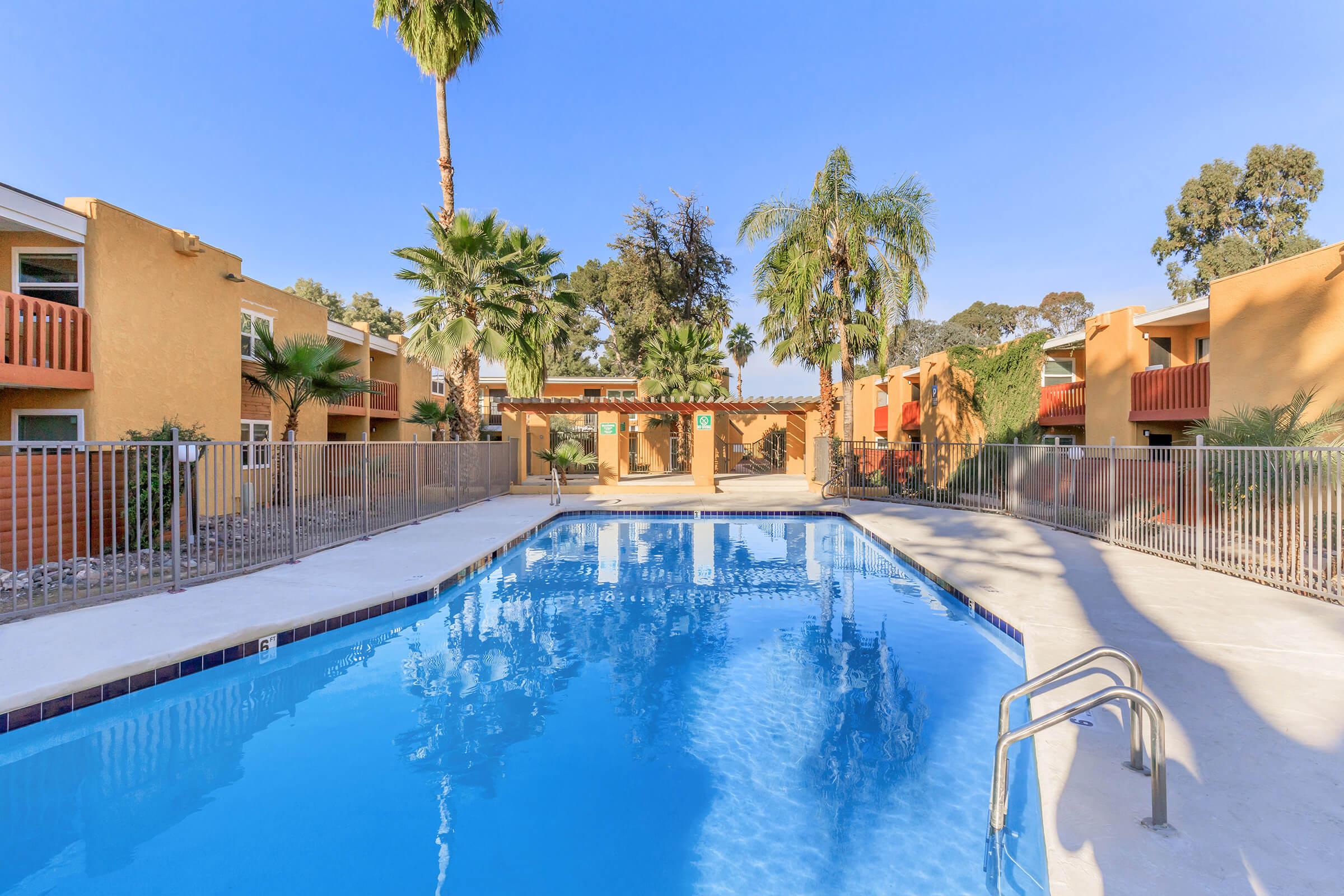 A swimming pool surrounded by palm trees and colorful apartment buildings. The clear blue water reflects the sky, and there are lounge areas and a gated entrance visible. The setting is bright and inviting, ideal for relaxation and leisure.