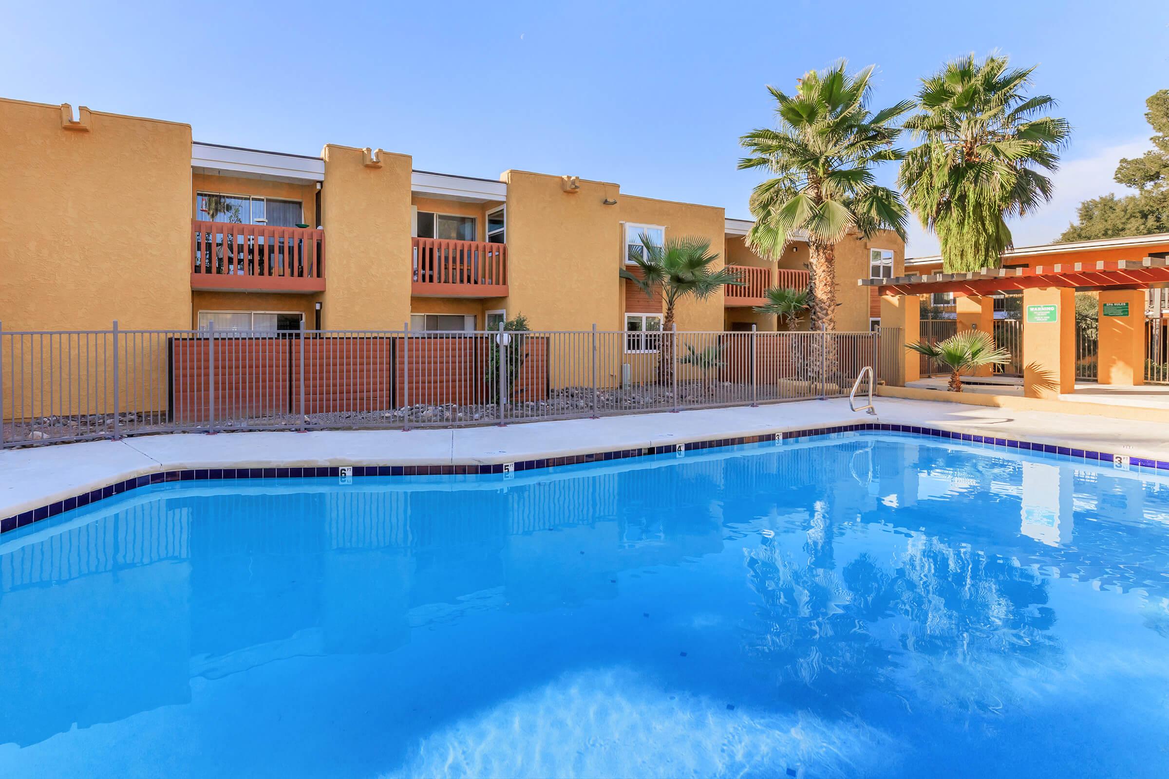 A clear blue swimming pool surrounded by a well-maintained area with palm trees and a fence. In the background, there are two-story buildings with balconies and orange walls, set against a bright blue sky. The scene conveys a relaxing outdoor atmosphere.