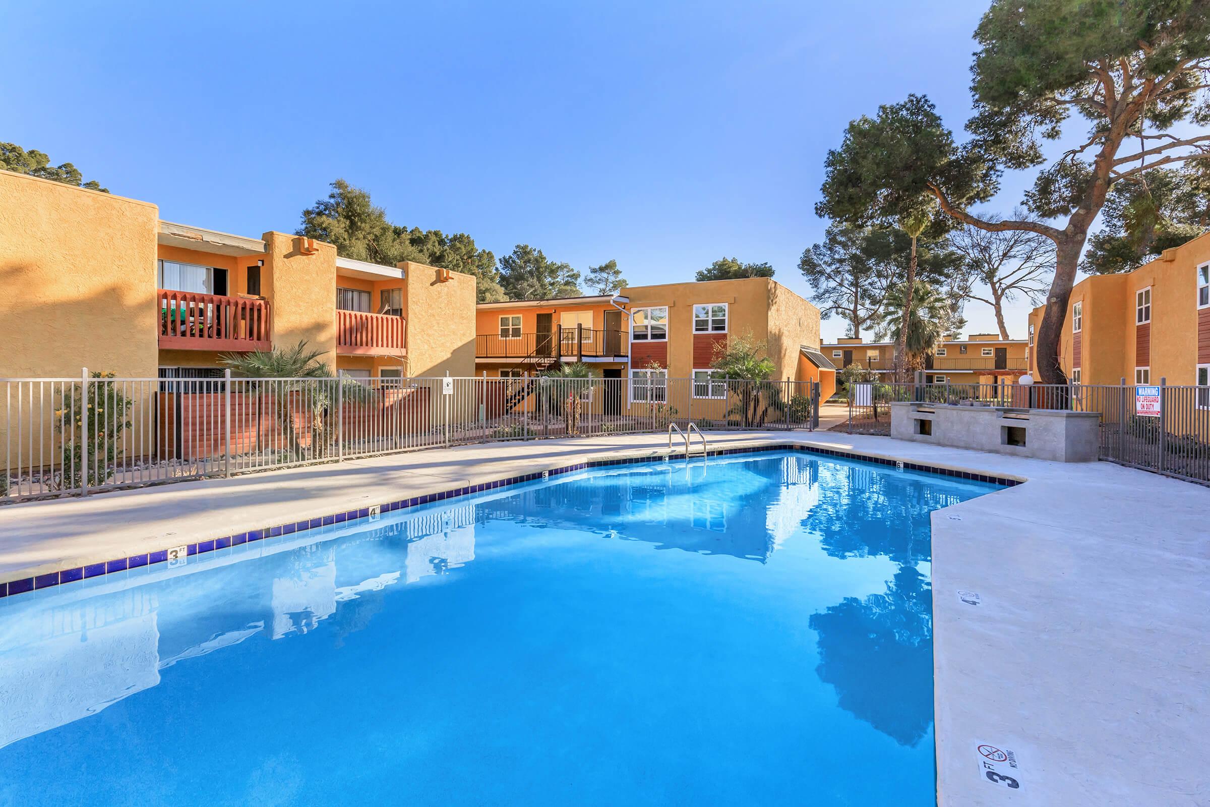 A clear blue swimming pool surrounded by a fence, with trees and two-story apartment buildings in the background. The buildings have a warm yellow exterior and balconies, creating a serene and inviting atmosphere. Bright sunlight enhances the vibrant colors of the scene.