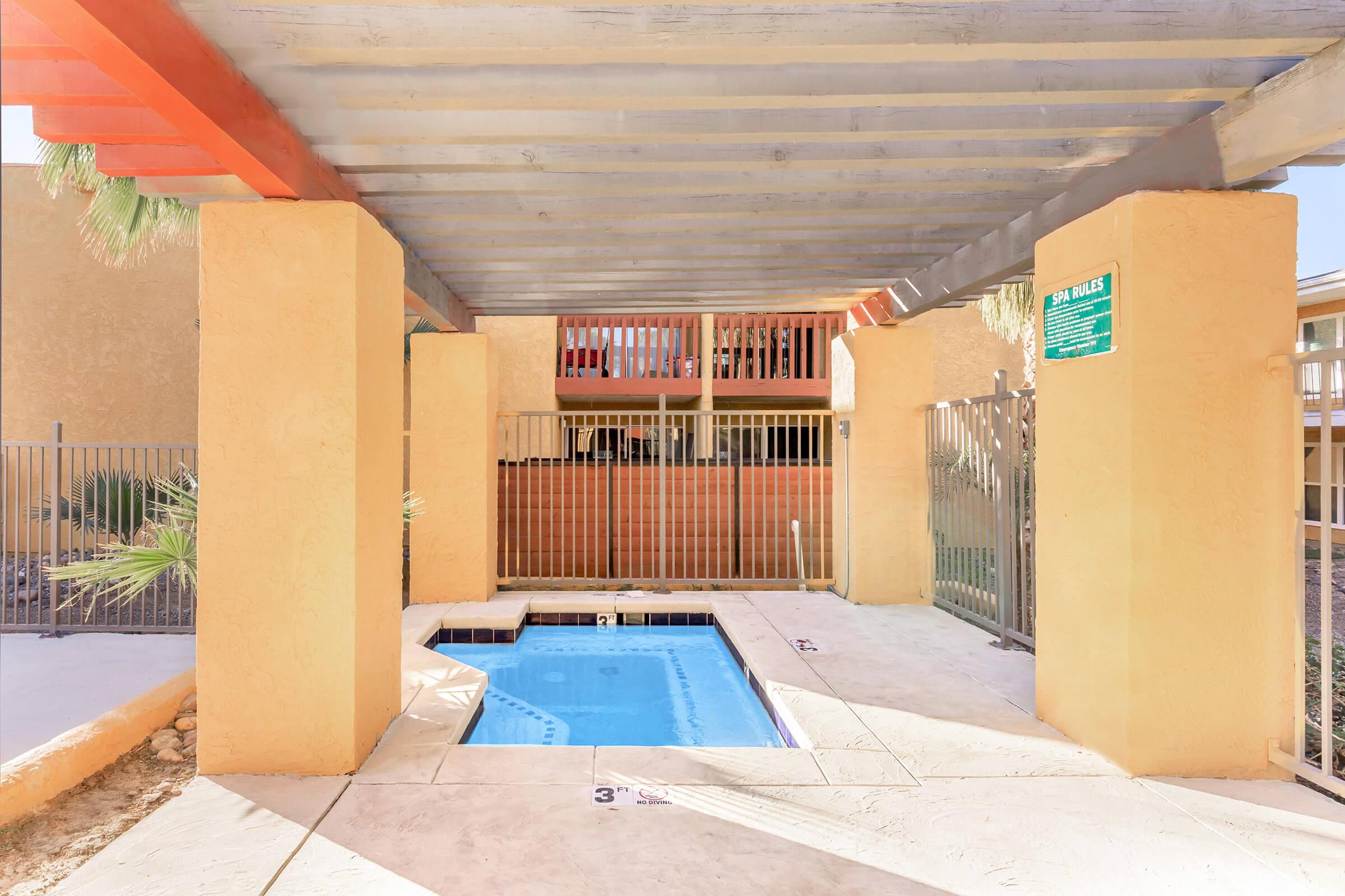 A small outdoor pool area with a covered structure. The pool is surrounded by a gated enclosure and features a shallow end marked with a "3 ft" indication. Palm trees and a wall are visible in the background, along with adjacent buildings providing a relaxed atmosphere.