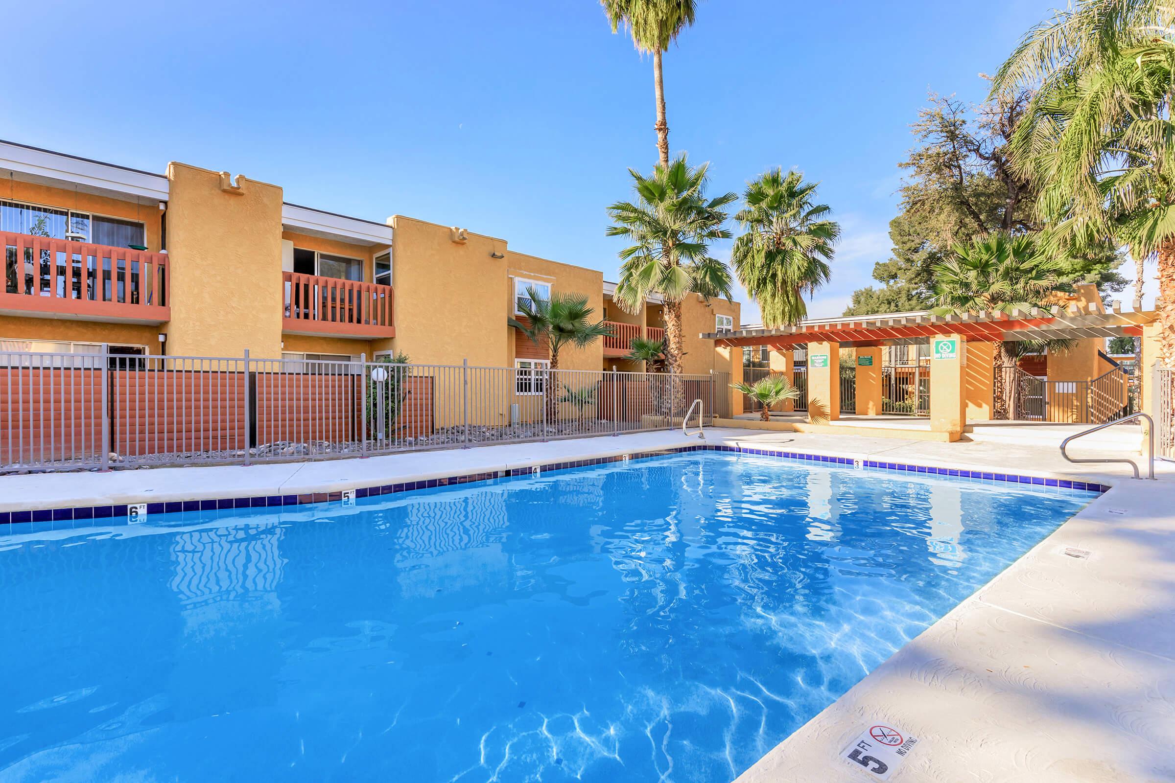 A sparkling blue swimming pool surrounded by palm trees, with an adjacent building featuring orange walls and balconies. A shaded seating area is visible in the background, enhancing the inviting atmosphere of the outdoor space. The clear sky suggests a sunny day.