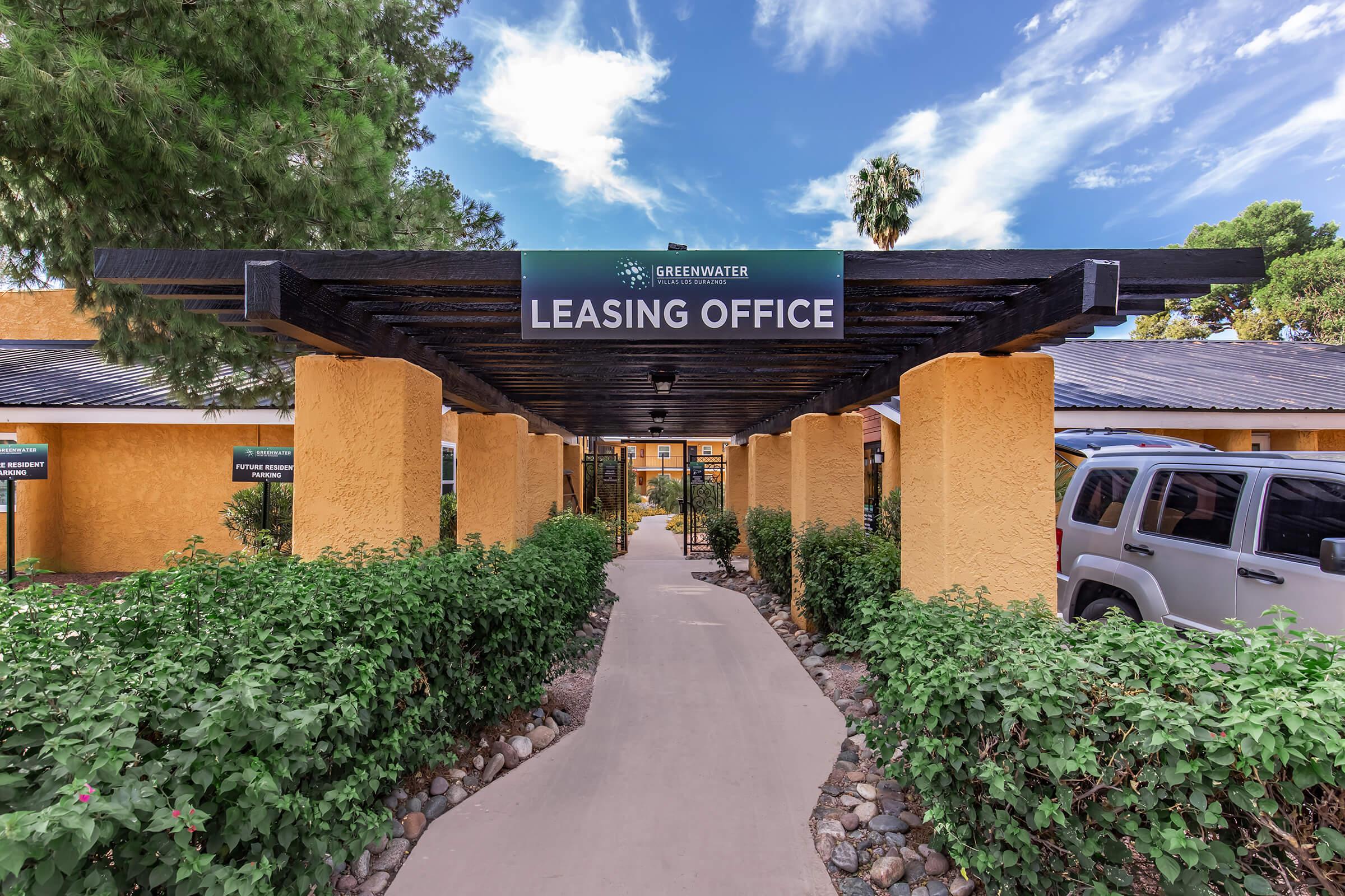 View of a leasing office exterior featuring a pathway lined with greenery and decorative stones. A prominent sign above reads "Leasing Office," and there are several palm trees in the background against a bright blue sky with some clouds. The surrounding buildings have a warm, yellow hue.