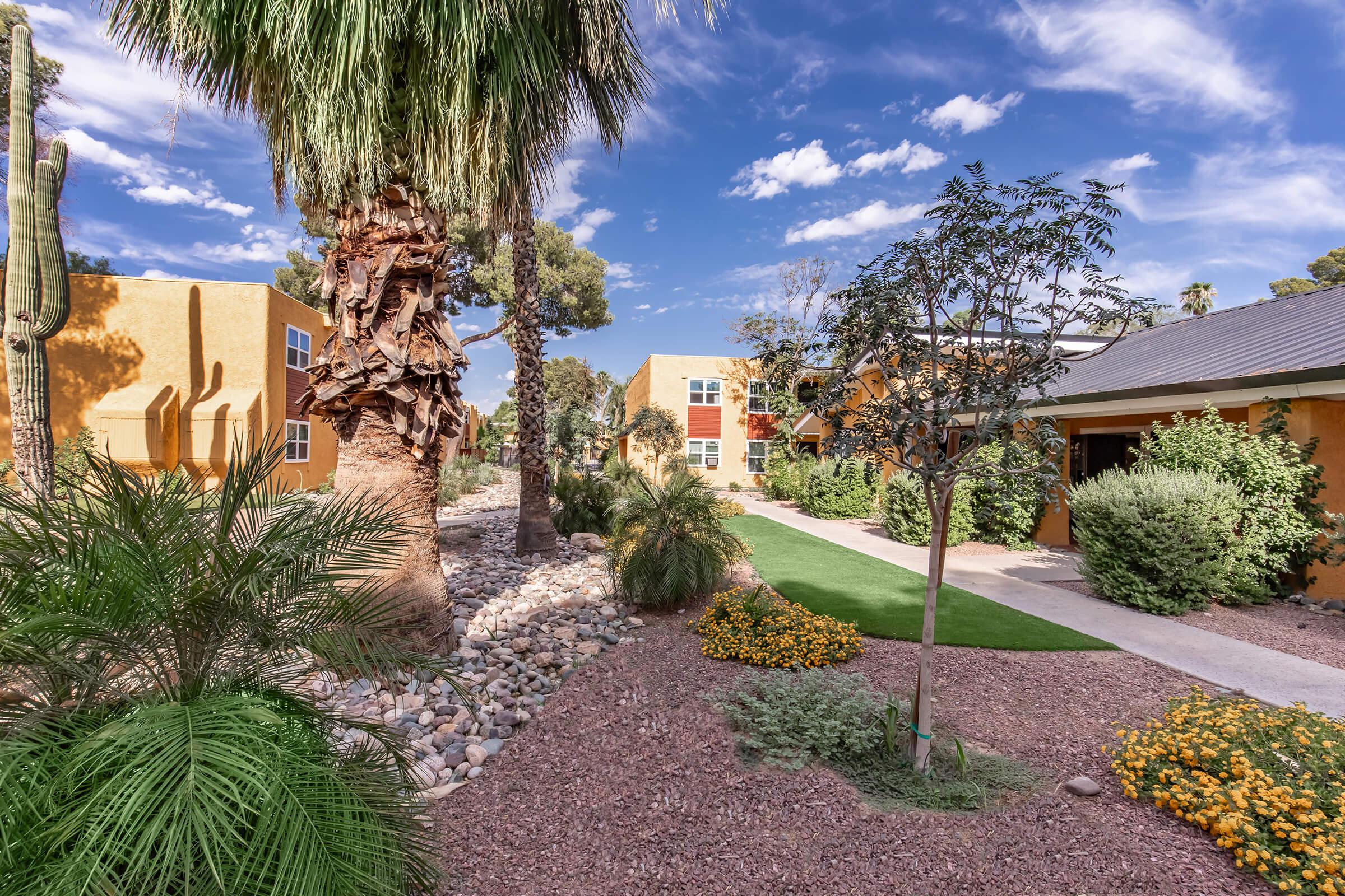 A landscaped pathway lined with palm trees and vibrant flowers, leading to residential buildings with colorful exteriors. The sky is partly cloudy, creating a bright and inviting atmosphere. The scene showcases a harmonious blend of greenery and architecture.