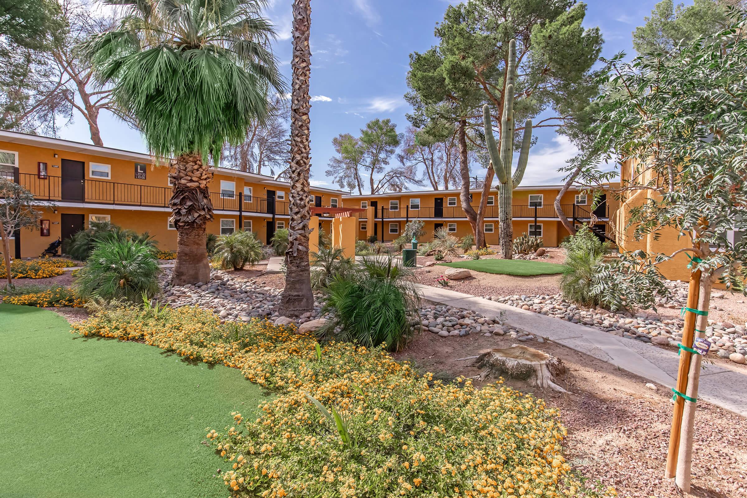 A lush courtyard scene featuring multiple yellow buildings surrounded by palm trees and colorful flower beds. A winding pathway leads through the greenery, creating a tropical and inviting atmosphere. The sky is partly cloudy, adding to the serene vibe of the setting.