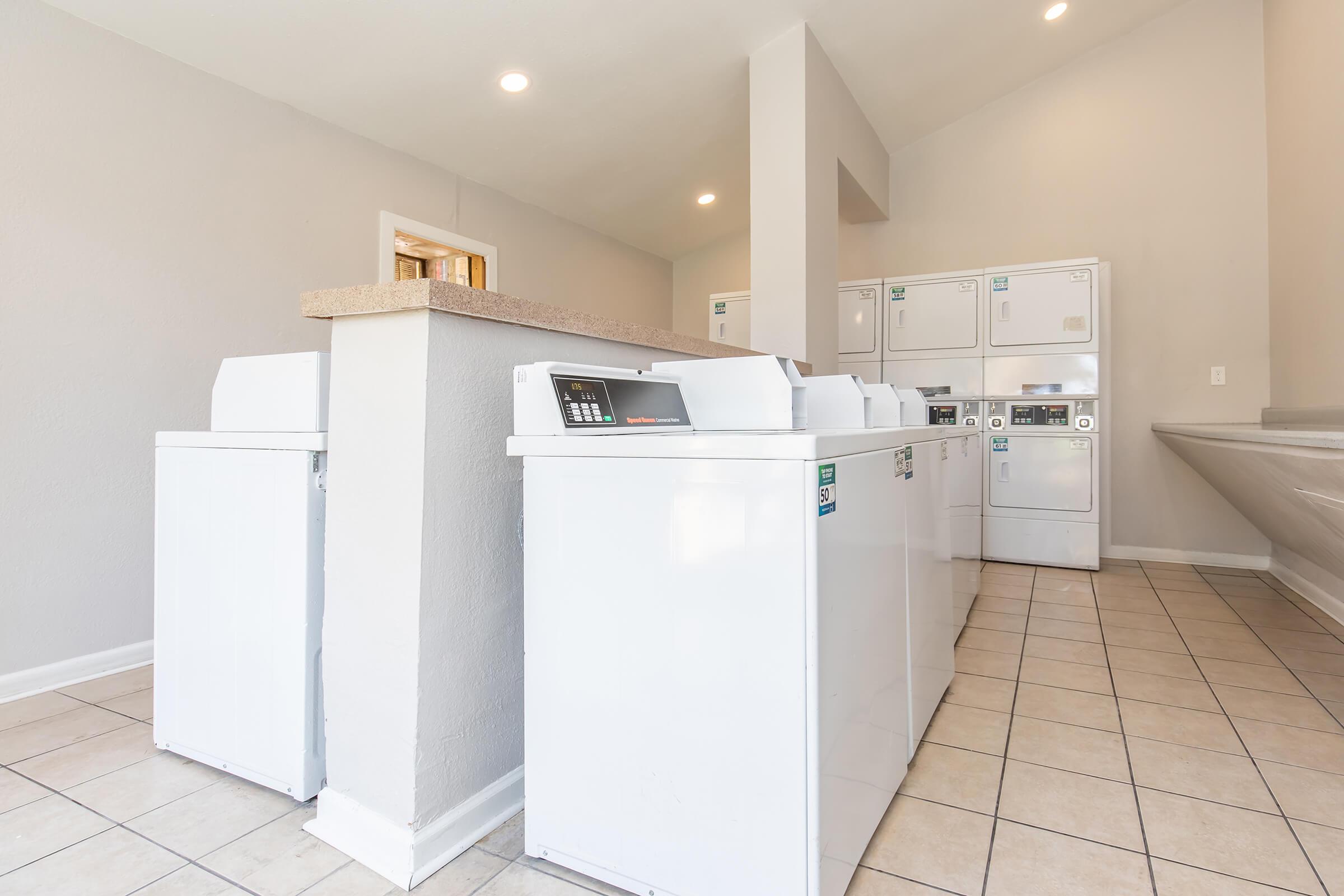 A clean and modern laundry room featuring several white washing machines and dryers lined up against the wall. The space has tiled flooring, bright lighting, and a counter area, creating a functional and inviting environment for doing laundry.