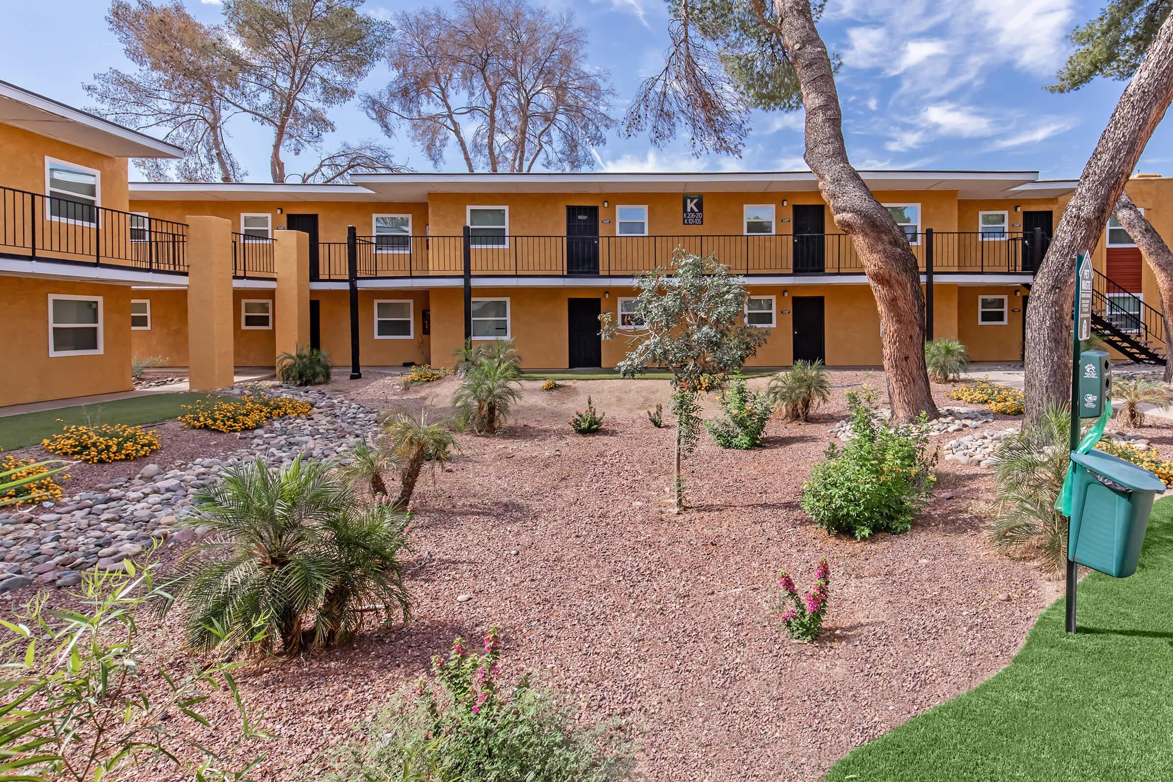 A brightly colored apartment complex featuring two-story buildings with balconies. The landscaping includes small shrubs, flower beds, and a small tree. The sky is partly cloudy, and there’s a green area with pathways and decorative rocks.