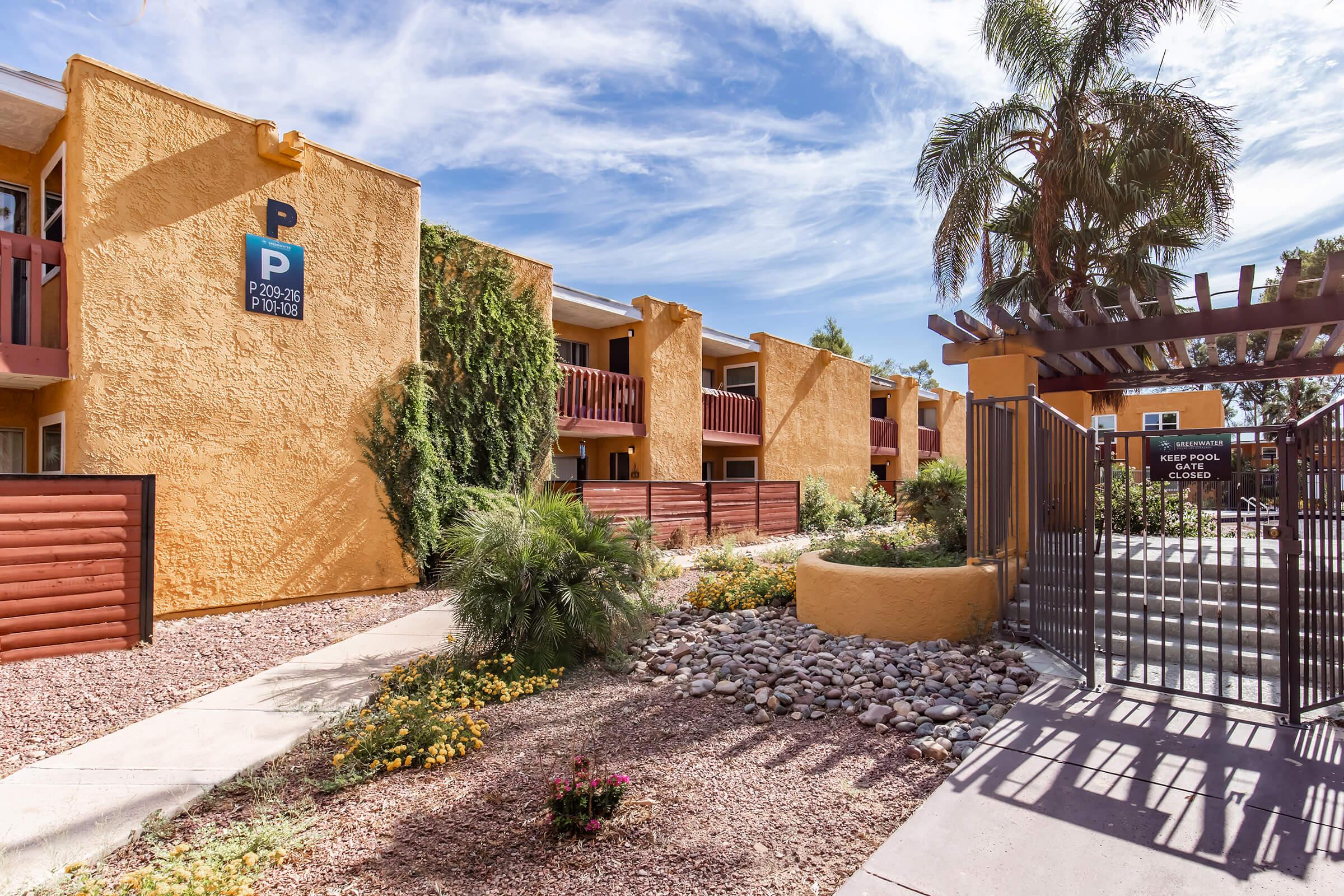 View of a bright yellow apartment complex with multiple units and balconies. A parking sign is visible alongside a landscaped area featuring palm trees and decorative rocks. A gated entrance leads into the complex, showcasing a clean and inviting environment under a partly cloudy sky.