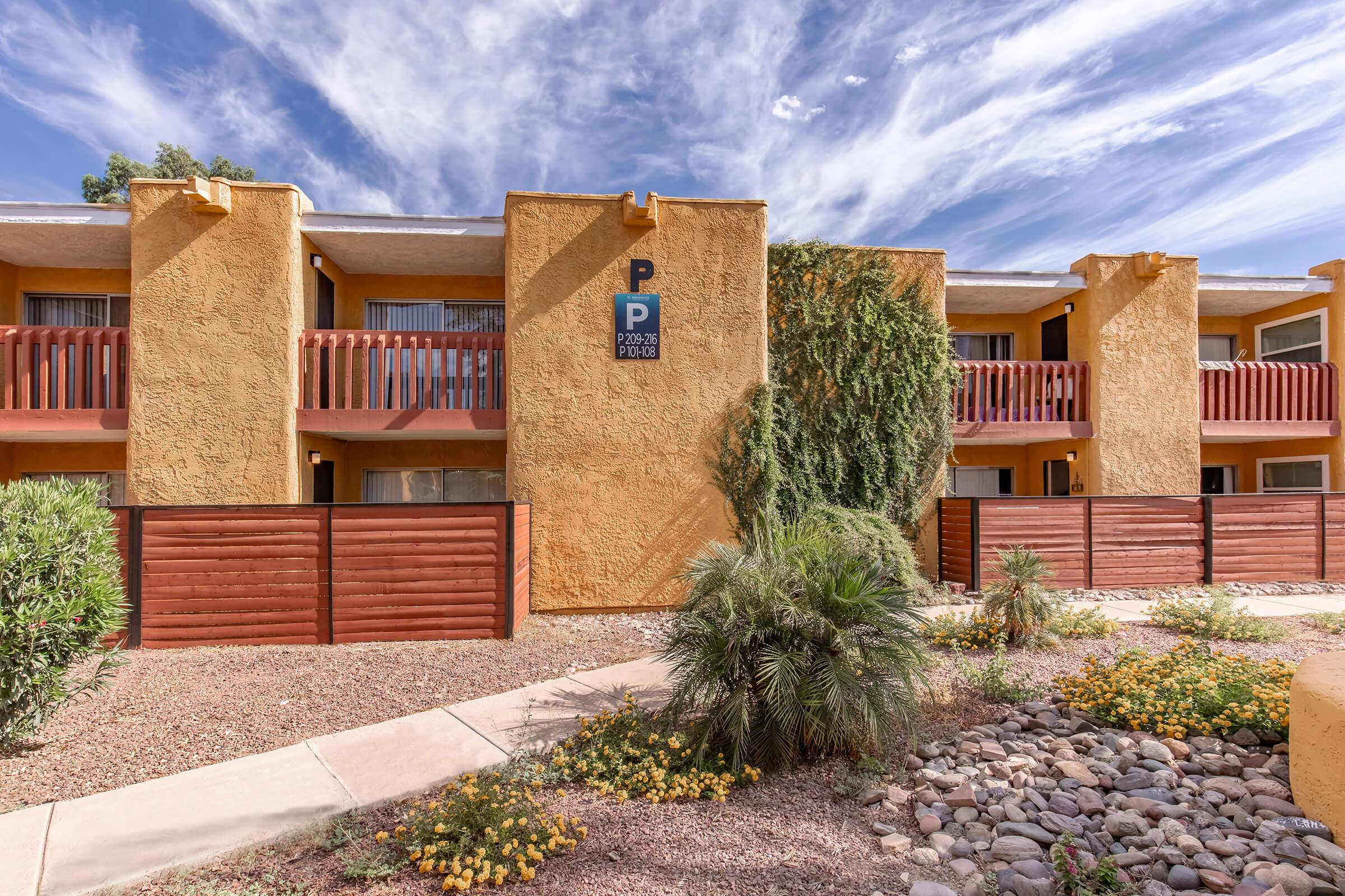 A view of a two-story building with a parking sign and balconies, painted in warm yellow tones. The surrounding landscape features low shrubs, a rock pathway, and desert plants under a bright blue sky with scattered clouds. The setting appears inviting and well-maintained.