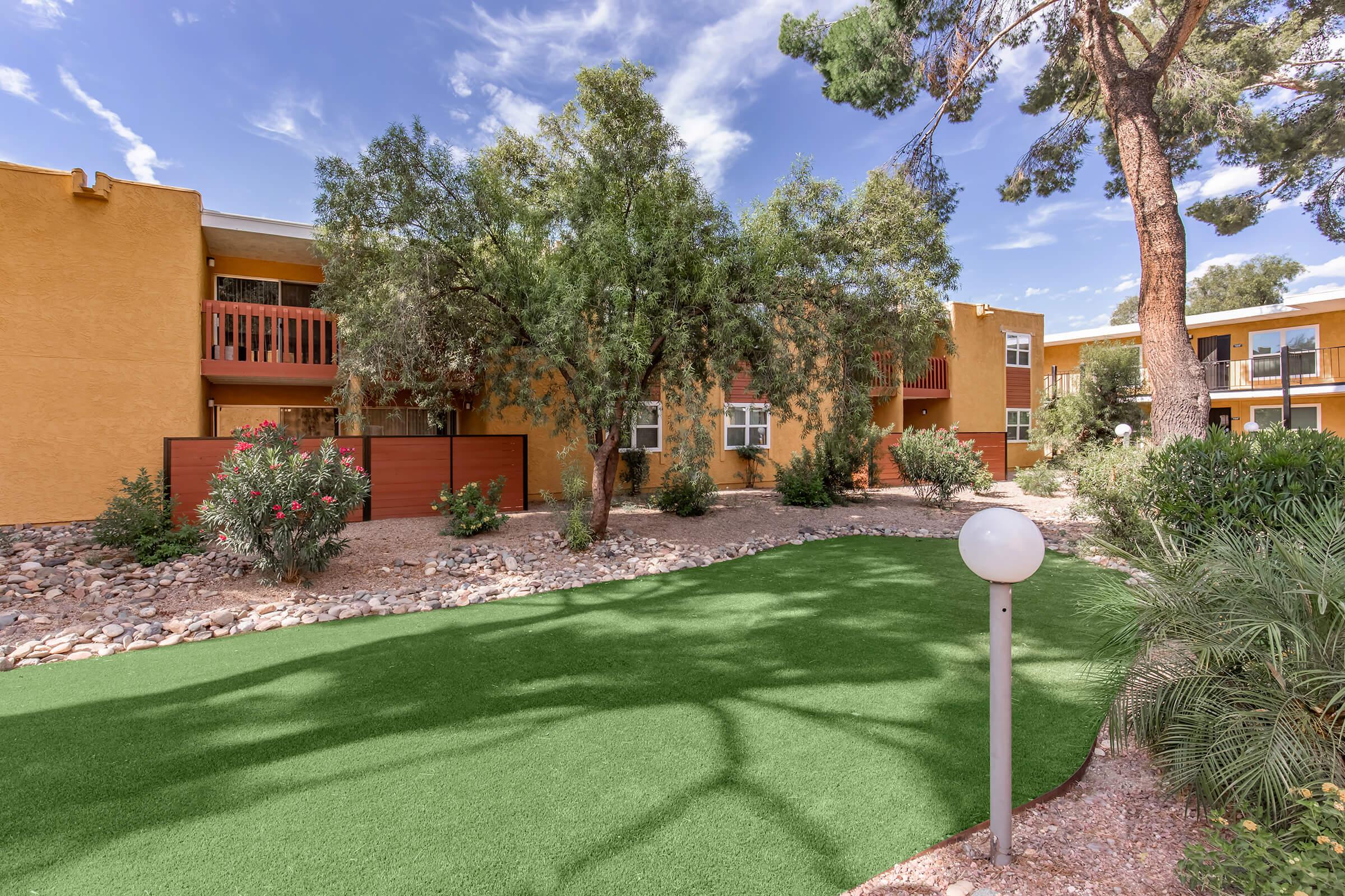 A well-maintained outdoor area with vibrant green artificial grass, surrounded by shrubs and trees. In the background, there are colorful apartment buildings with balconies. A round lamp post is located in the foreground, adding to the aesthetic of the landscaped space. Blue sky with scattered clouds is visible above.