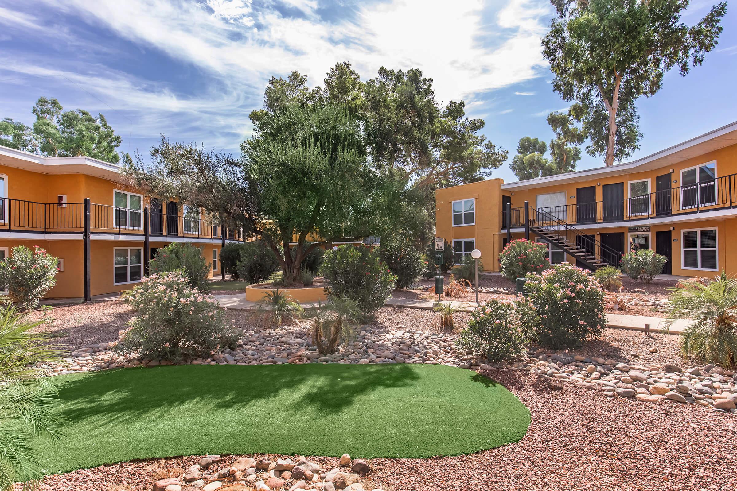 A landscaped courtyard featuring several two-story yellow buildings with black railings. The area includes shrubs and small trees, with a patch of artificial grass in the foreground. The sky is partly cloudy, creating a bright and inviting atmosphere.