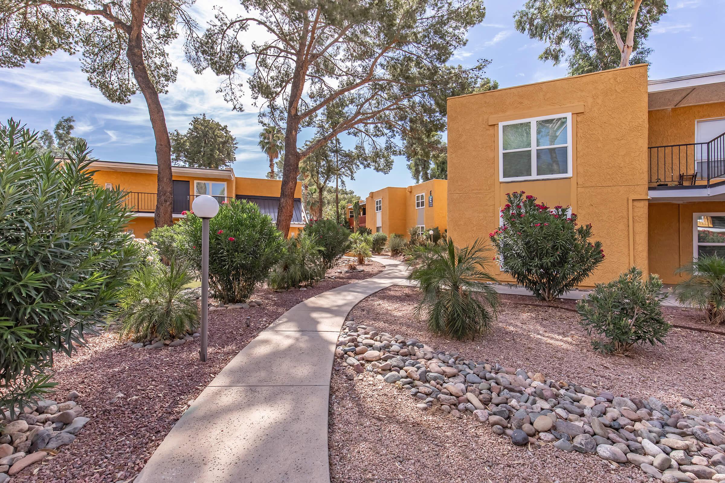 A landscaped path winds through a courtyard surrounded by yellow apartment buildings. Lush green trees and shrubs line the pathway, which is adorned with smooth stones. The sky is clear with some clouds, creating a serene outdoor setting.