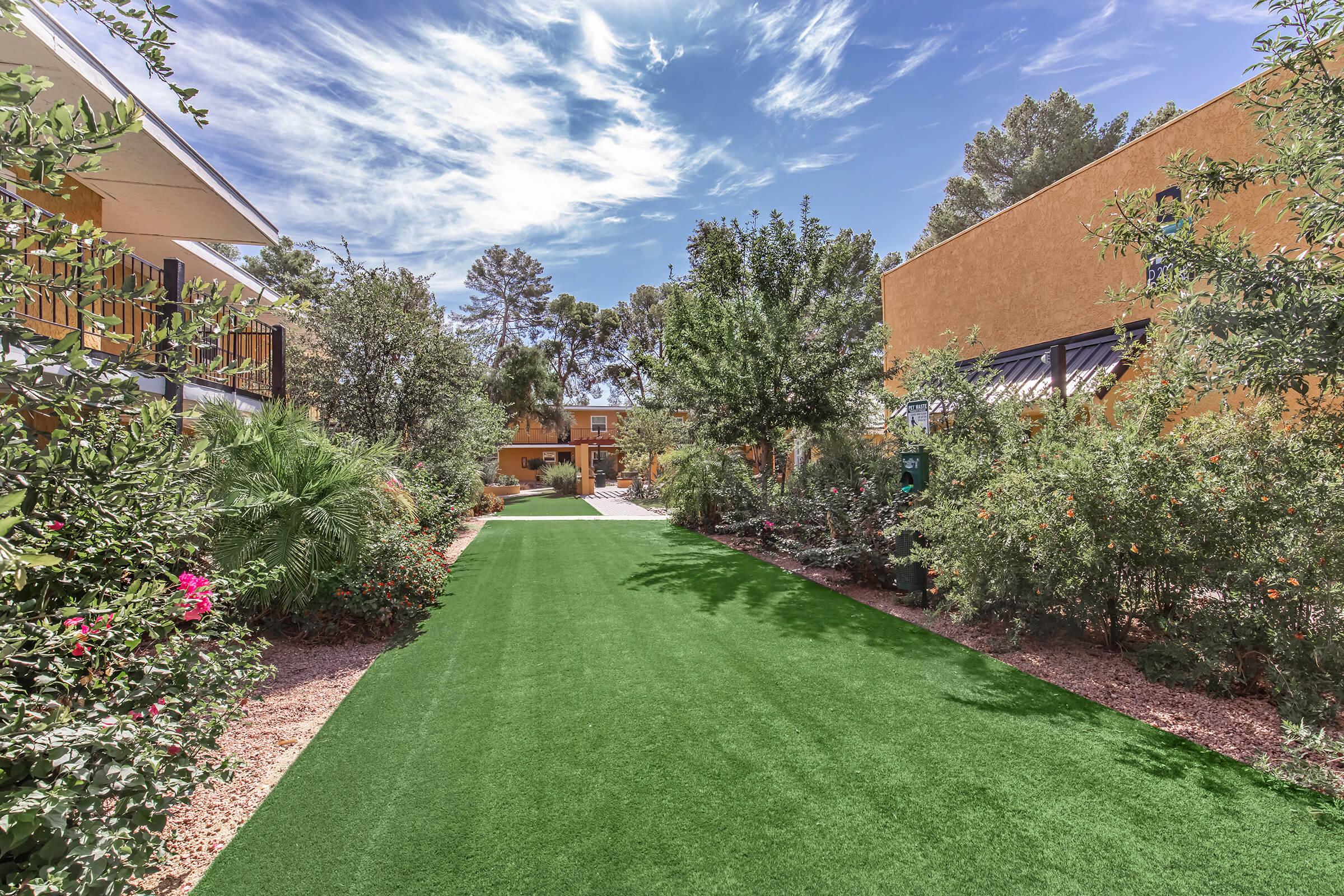A tranquil pathway through a landscaped area featuring vibrant green artificial grass, flanked by lush shrubs and flowering plants. Bright blue sky with scattered clouds above, and buildings with warm, earthy tones visible in the background, creating a peaceful outdoor atmosphere.