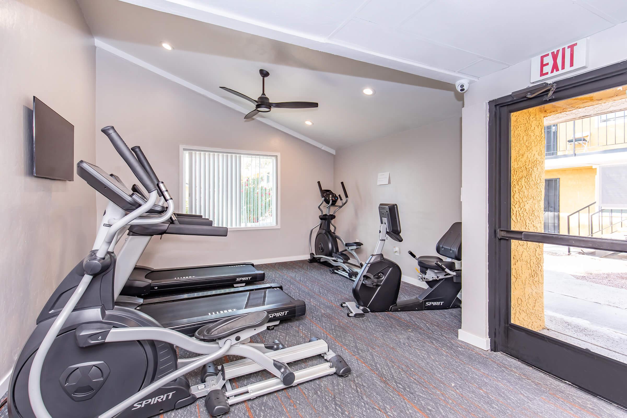 A fitness room featuring exercise equipment, including a treadmill, elliptical machine, and stationary bike. The space is well-lit, with a ceiling fan and a television mounted on the wall. A door on the right leads outside, and there are large windows for natural light.