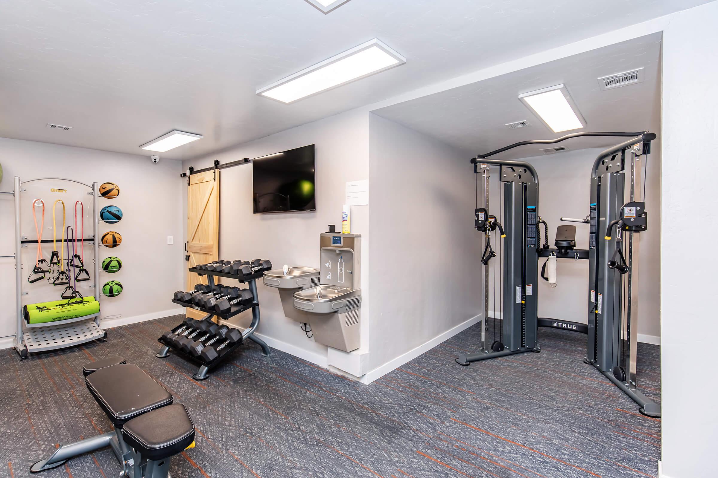 A modern fitness center featuring weightlifting equipment, a multi-gym, and a water fountain. The space is well-lit with overhead lights, and there is a TV mounted on the wall. Rubber exercise mats and various exercise accessories are visible, providing a comprehensive workout environment.