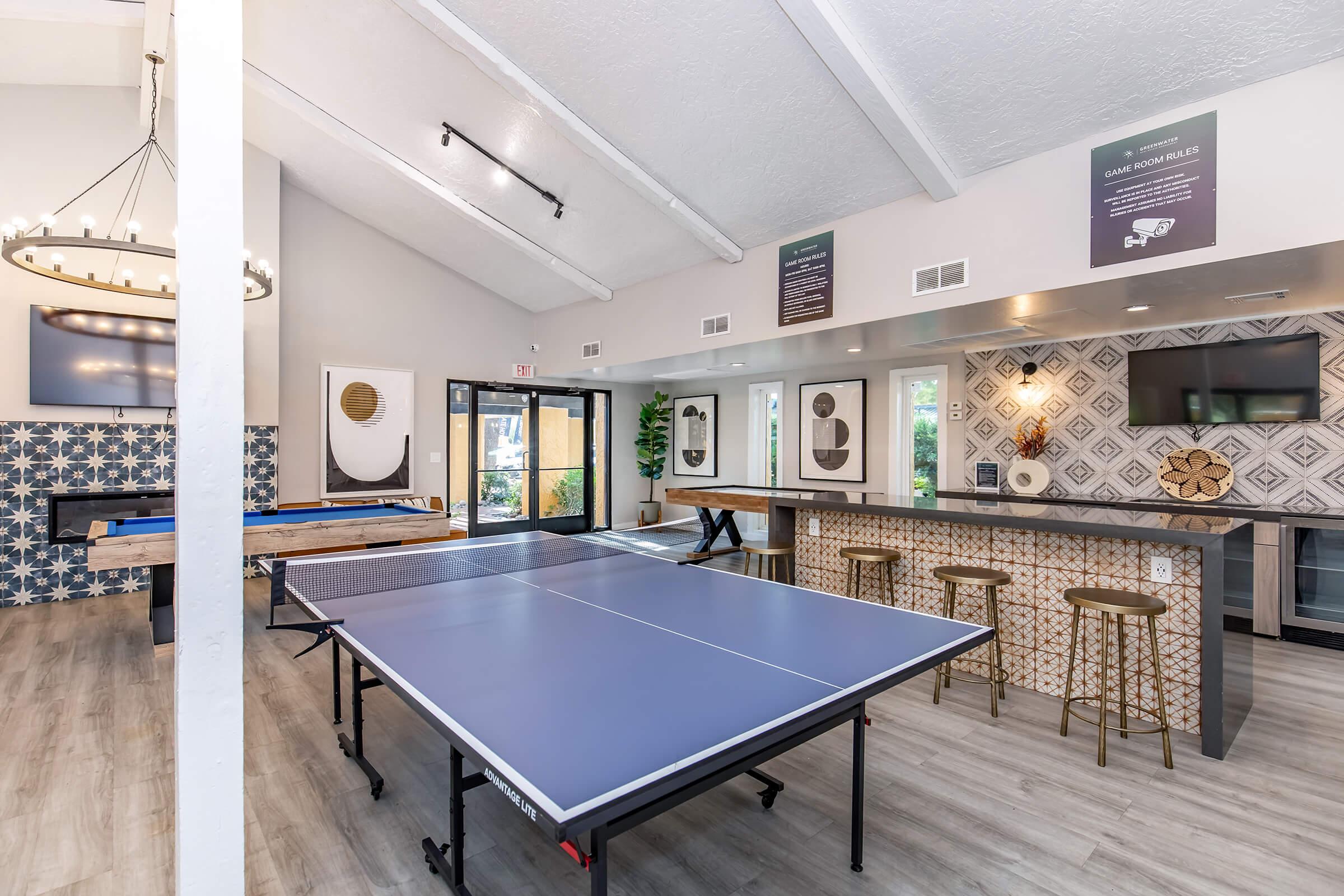 Interior view of a recreational room featuring a blue ping pong table, a bar area with high stools, and decorative wall art. The space has high ceilings, large windows for natural light, and a stylish patterned backsplash. Additional game tables are visible, creating a fun and inviting atmosphere.