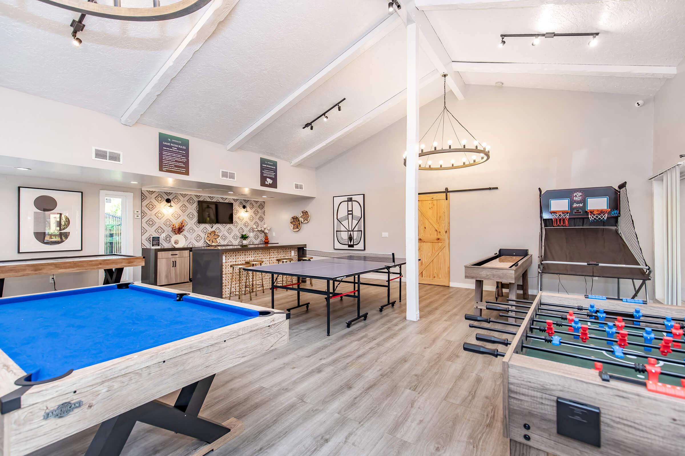 A spacious game room featuring various leisure activities, including a blue pool table, a ping pong table, foosball table, and a basketball shooting game. The room has high ceilings, stylish decor, and a modern design, with wood flooring and a bar area in the background.