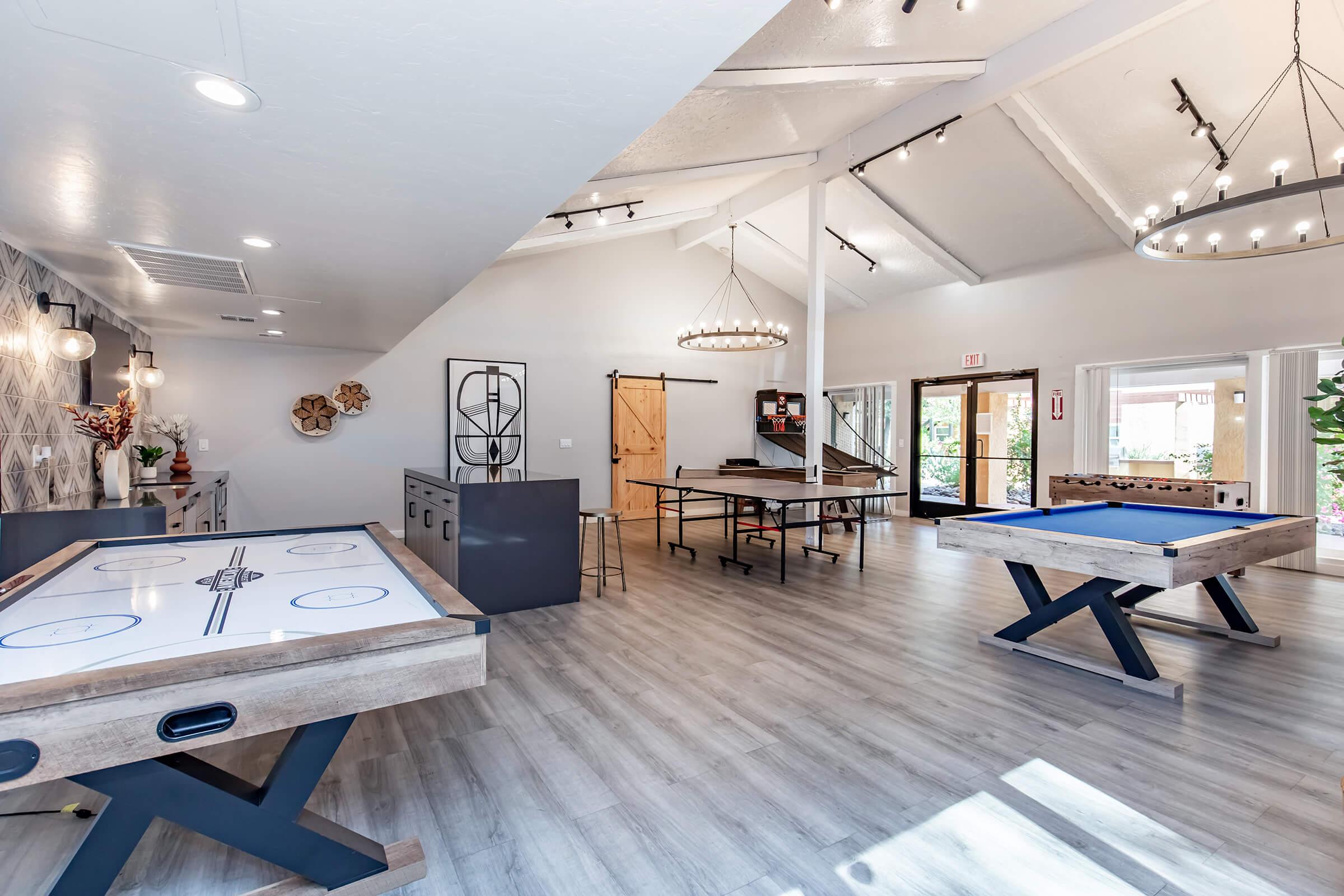 Interior of a recreational room featuring a pool table, air hockey table, and a ping pong table. The space has a modern design with high ceilings, pendant lighting, and large windows allowing natural light. Decorative elements include wall art and plants, creating a welcoming atmosphere for socializing and entertainment.