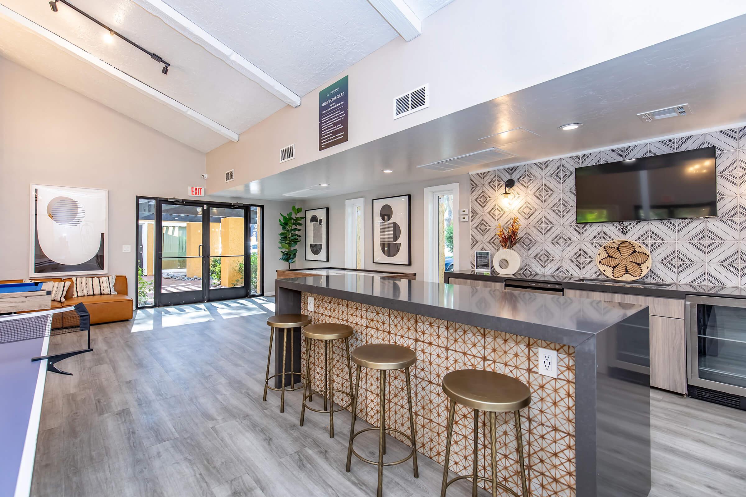 Modern interior of a welcoming communal space featuring a sleek kitchen island with four gold bar stools, geometric patterned backsplash, large screen TV mounted on the wall, and seating area with contemporary art. Large windows provide natural light.