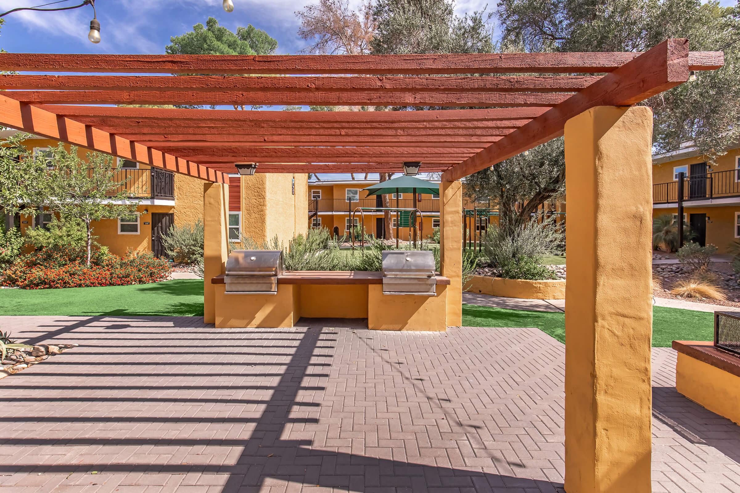 Outdoor barbecue area featuring two grills under a wooden pergola, surrounded by lush greenery and colorful landscaping. The ground is paved with bricks, and there is a shaded seating area with an umbrella in the background, creating a welcoming space for gatherings and meals.